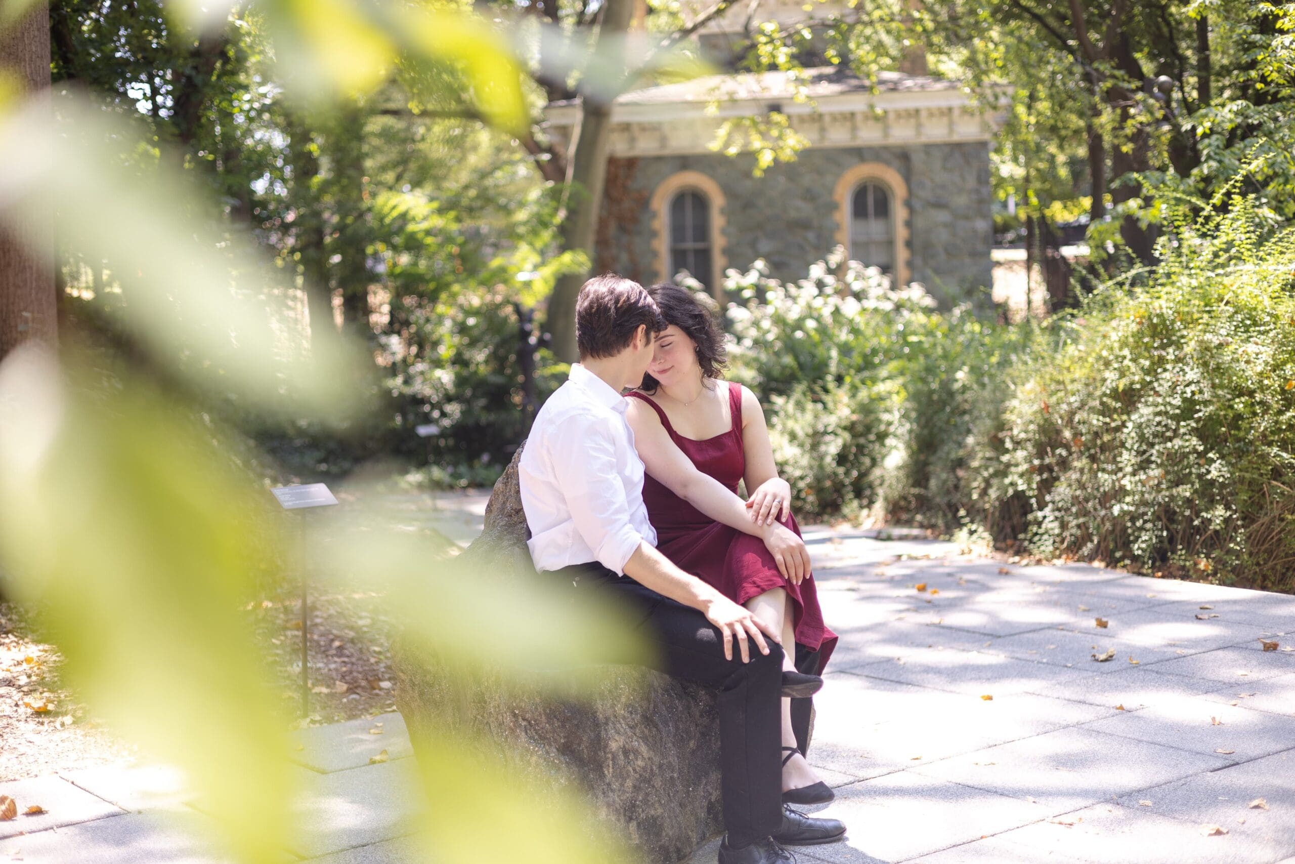 A couple sits closely on a rock in The Baltimore Museum of Art garden, surrounded by trees. They gaze affectionately at each other, creating a serene, romantic atmosphere.