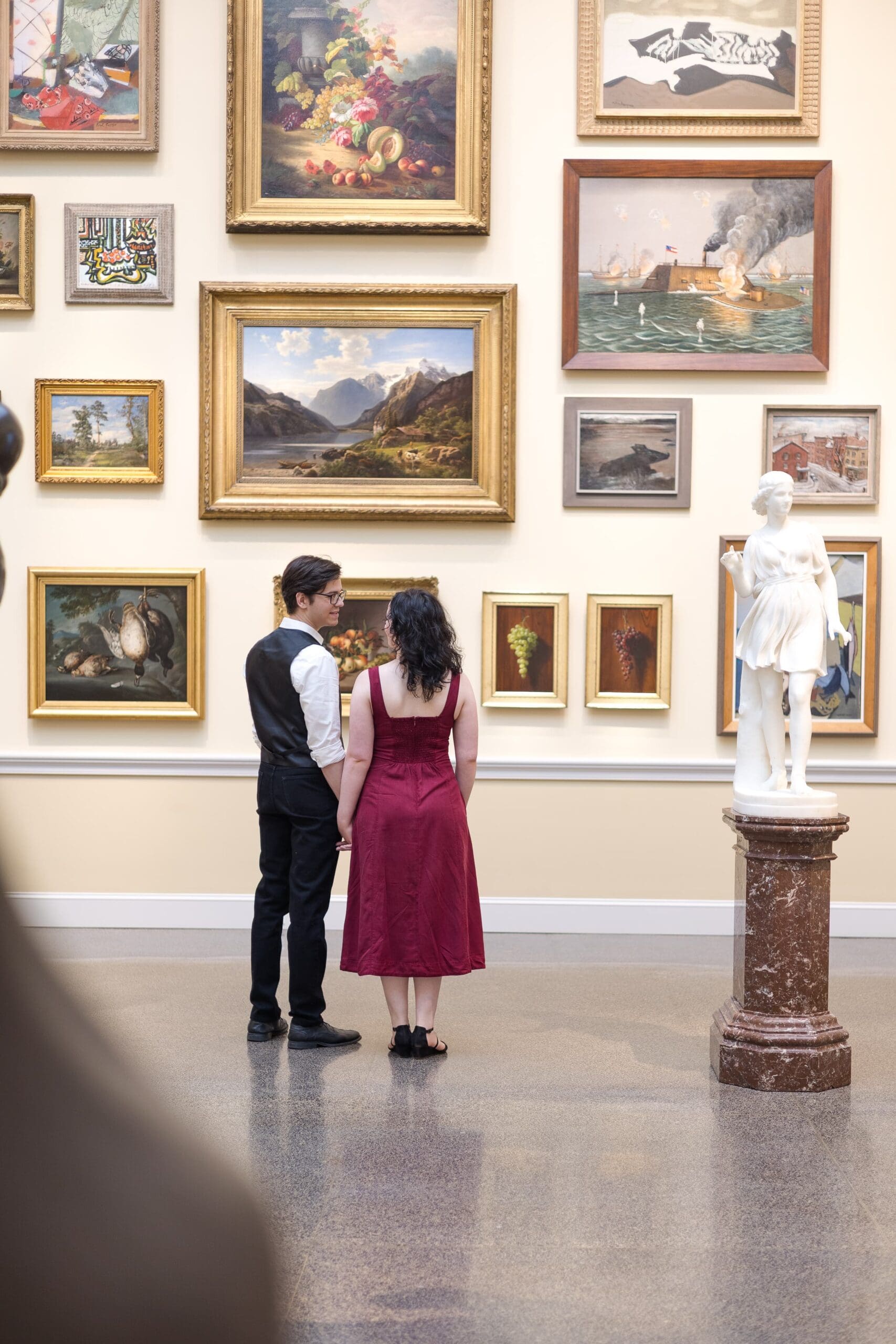 Couple hold each other close in an art exhibit. Surrounded by art hanging on the wall, but they can only look at each other.