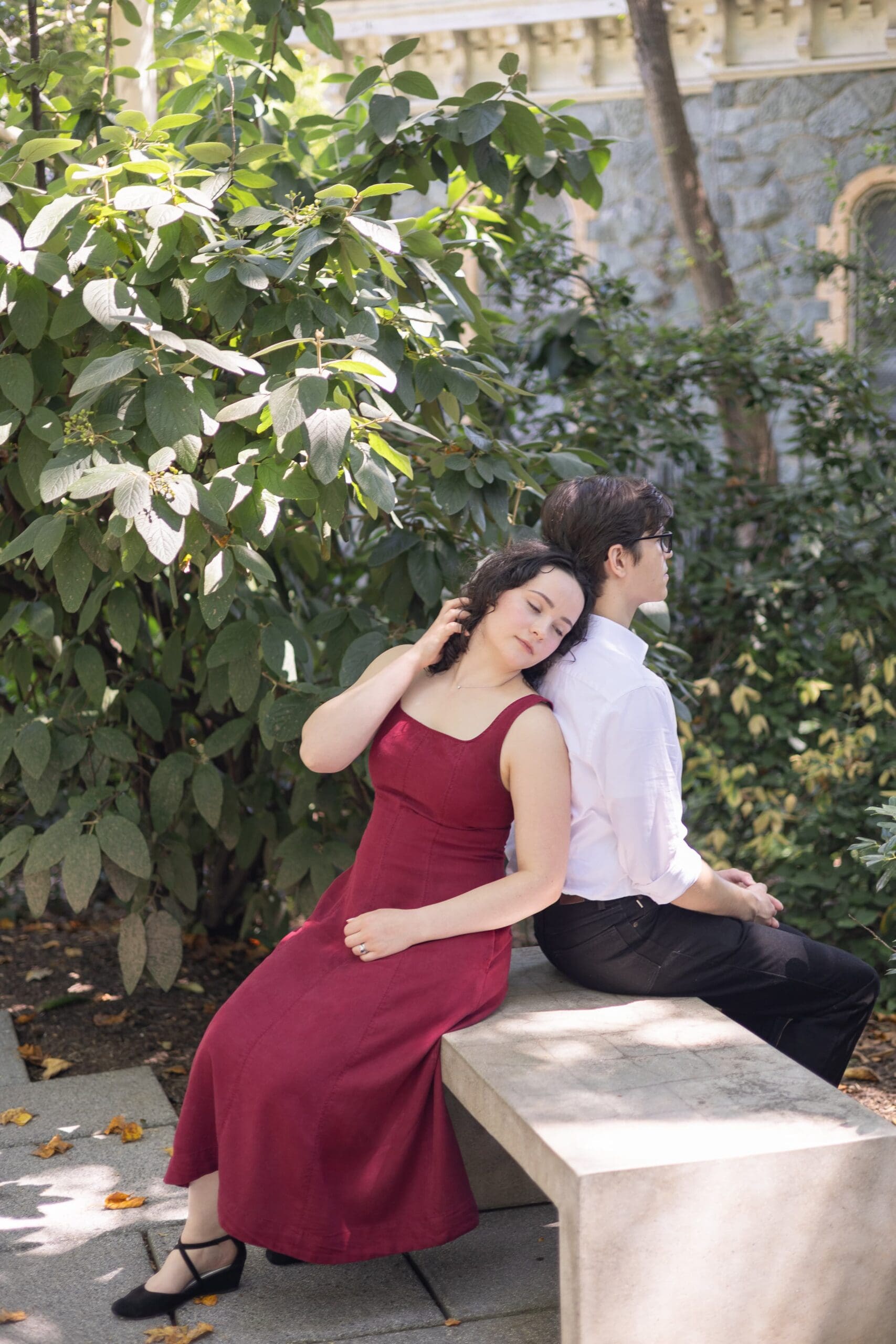 A couple sits closely on a bench in The Baltimore Museum of Art garden, surrounded by trees. The woman plays with her hair affectionately, while the man looks off into the distance creating a serene, romantic atmosphere.