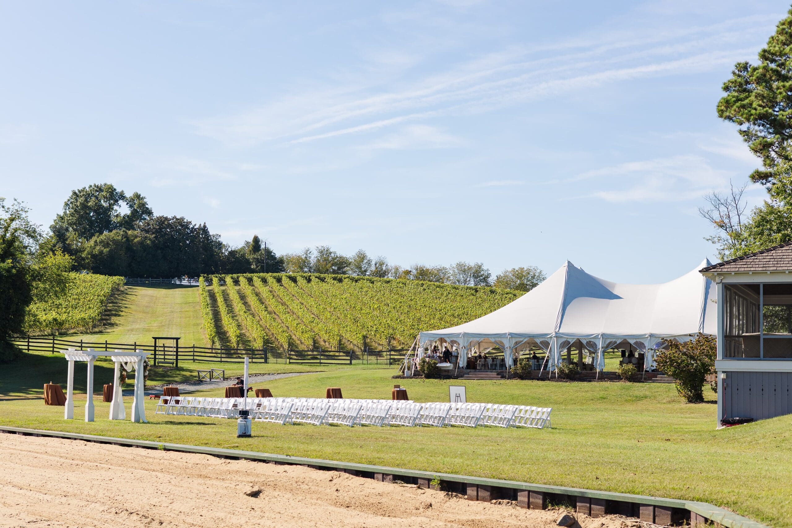 Wedding venue in a vineyard with a large white tent and rows of white chairs on a green lawn. The sky is clear, creating a serene, elegant setting.