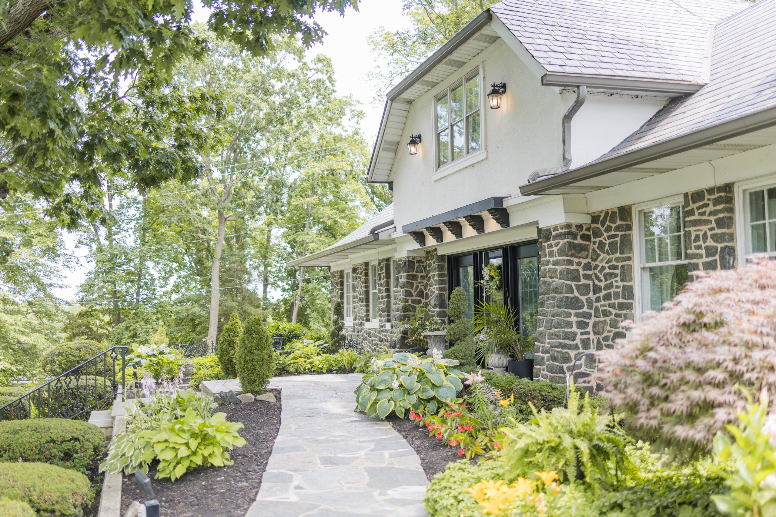 A stone and stucco house with a landscaped front yard featuring various plants, flowers, and a stone path leading to the entrance.