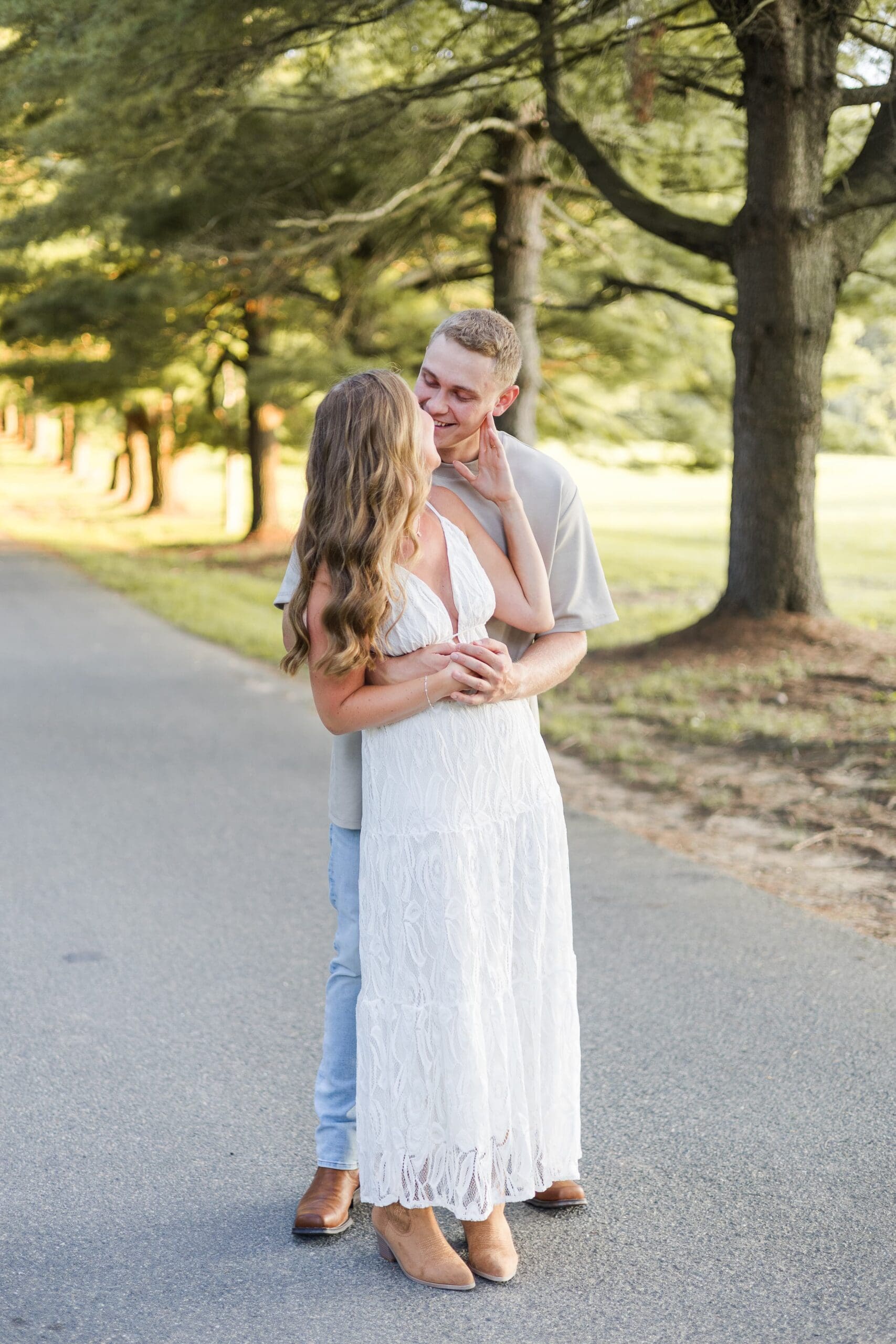 A couple stands on a tree-lined pavement. The woman in a white dress and the man in a cream shirt nuzzle together holding one another, smiling.