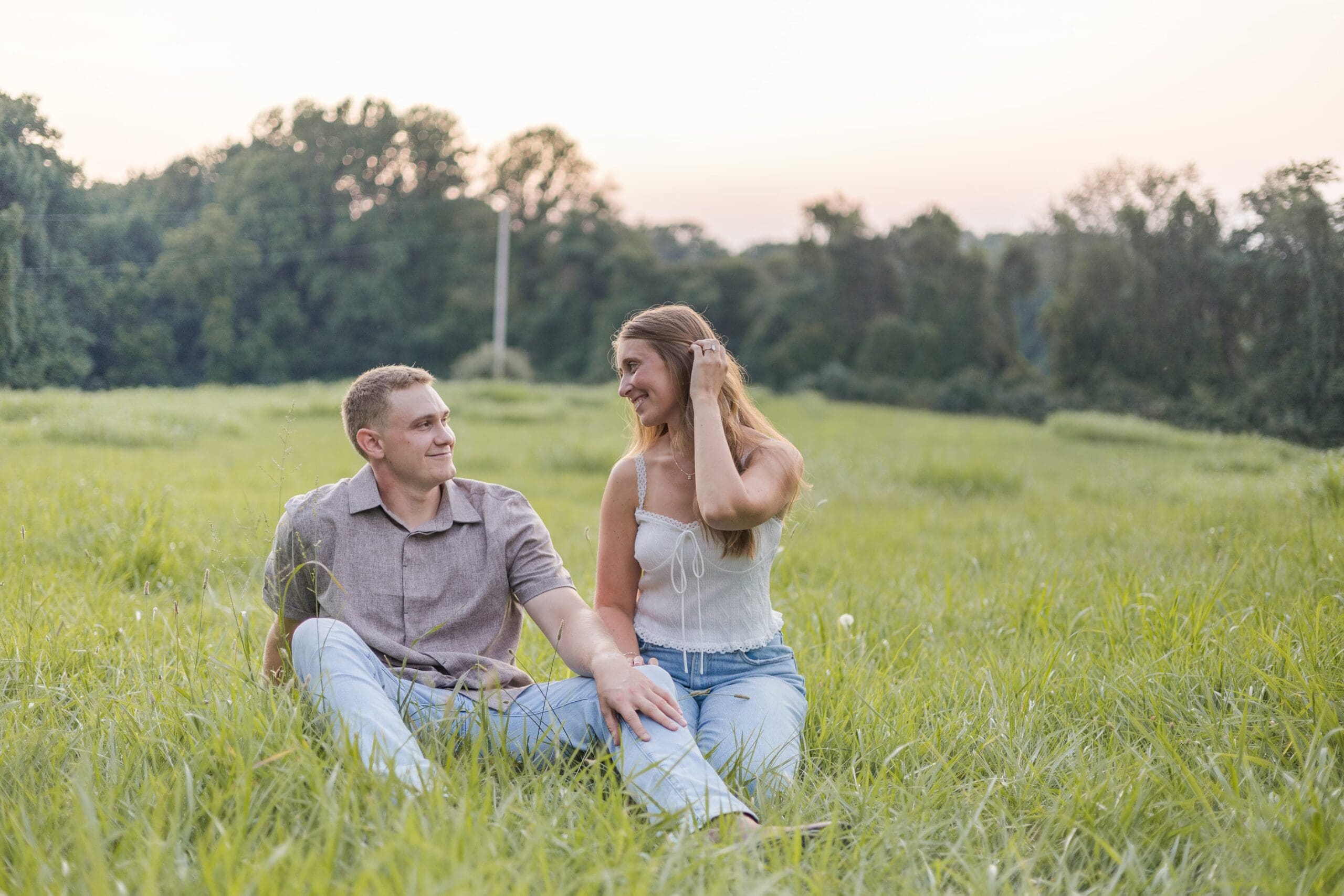 A couple sits in a grassy field at sunset, smiling at each other. The man wears a patterned shirt, and the woman is in a tank top and jeans. Trees are in the background.
