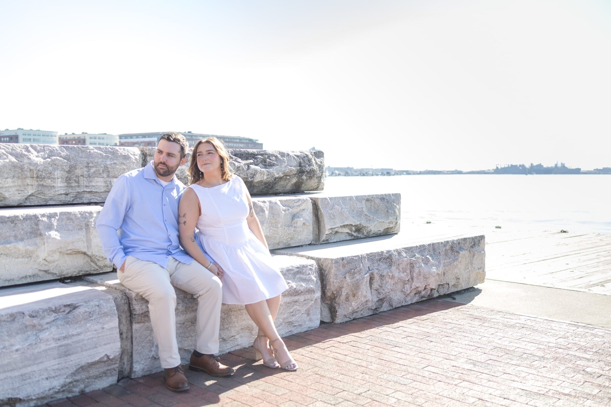 Couple sits on a stone staircase in Fells Point, Baltimore. The sun shiines while the calm waters are behind them.