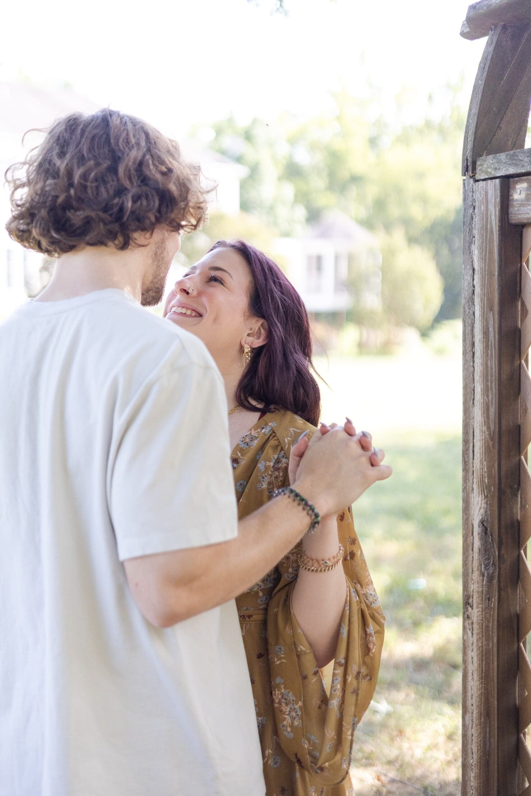 A couple holds hands and smiles lovingly at each other in a sunlit garden. The background is blurred, emphasizing their joyful connection and warmth.