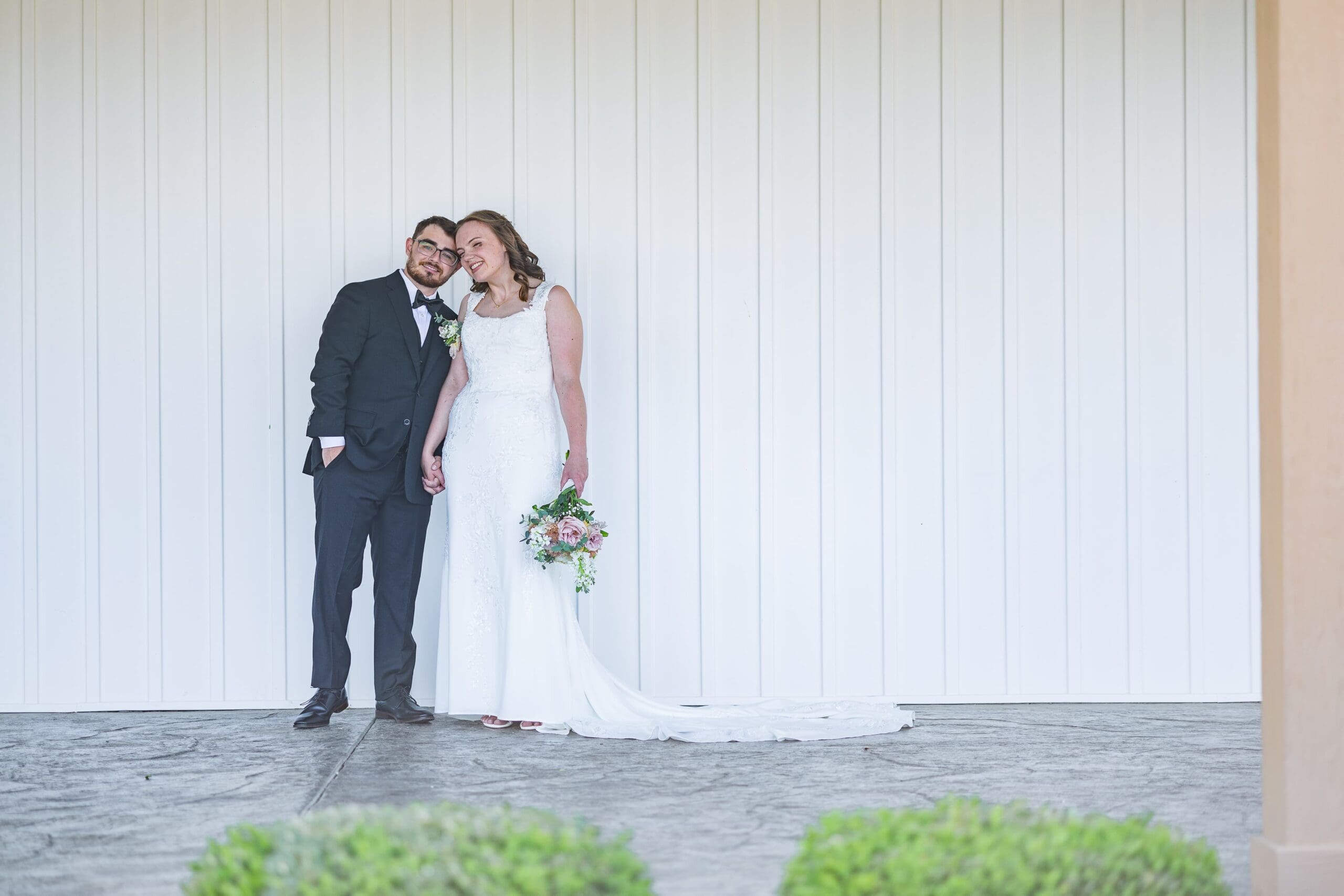 A smiling bride and groom stand closely together in front of a white paneled wall. She holds a bouquet; both exude joy and elegance.