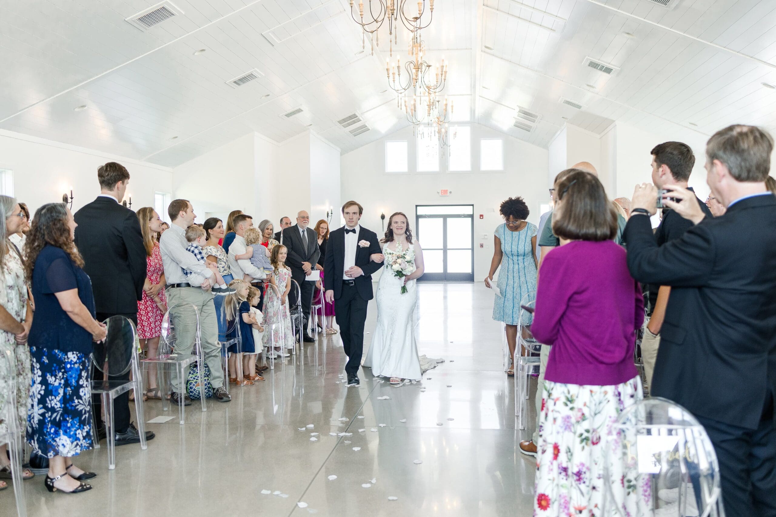 A bride in a white dress walks down the aisle of a bright, spacious hall filled with seated guests. People stand, smiling and capturing the moment.