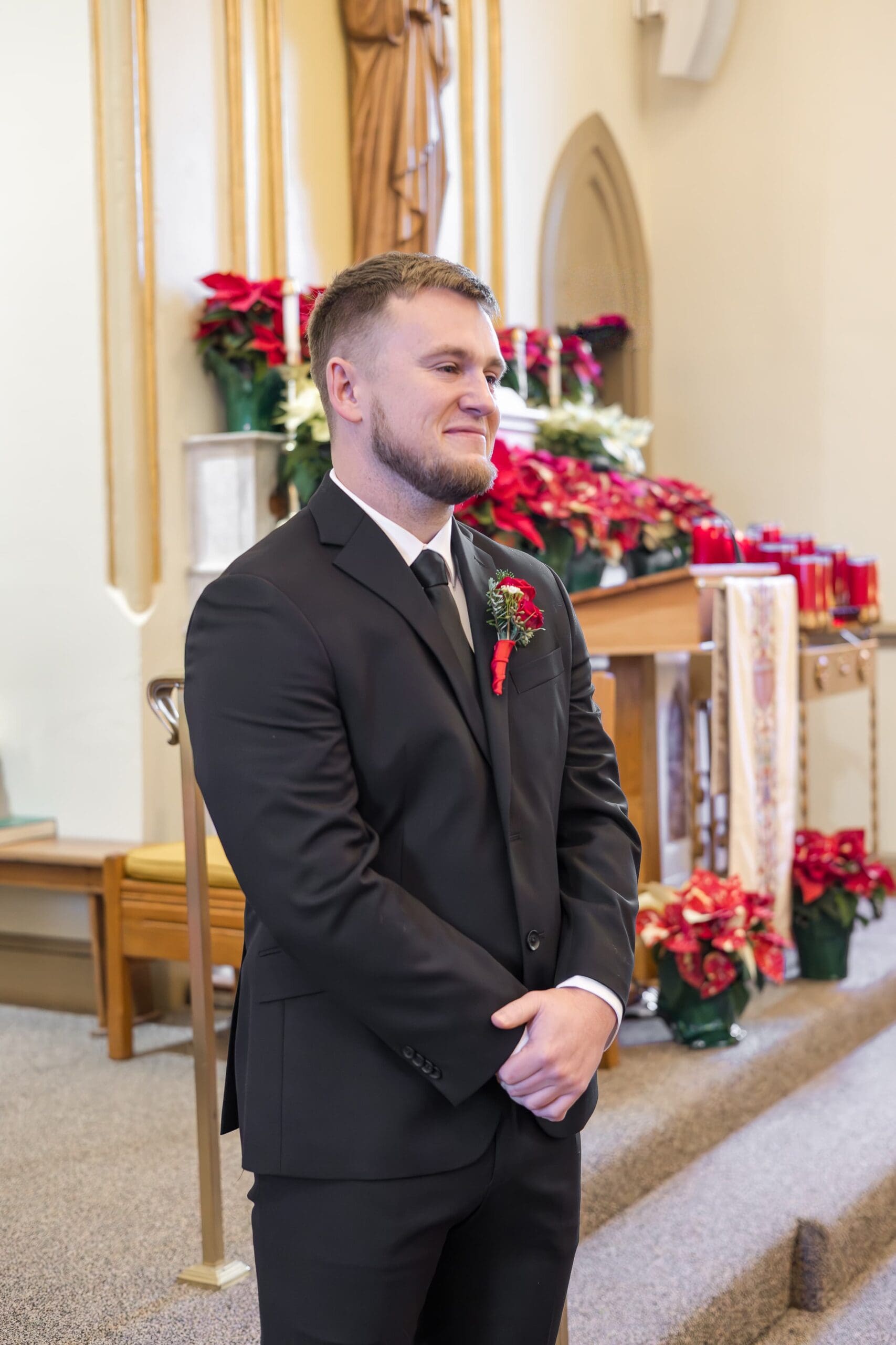 man at the alter stands waiting for his bride to meet him at the altar, smiling at her in a black suit and red boutonniere.