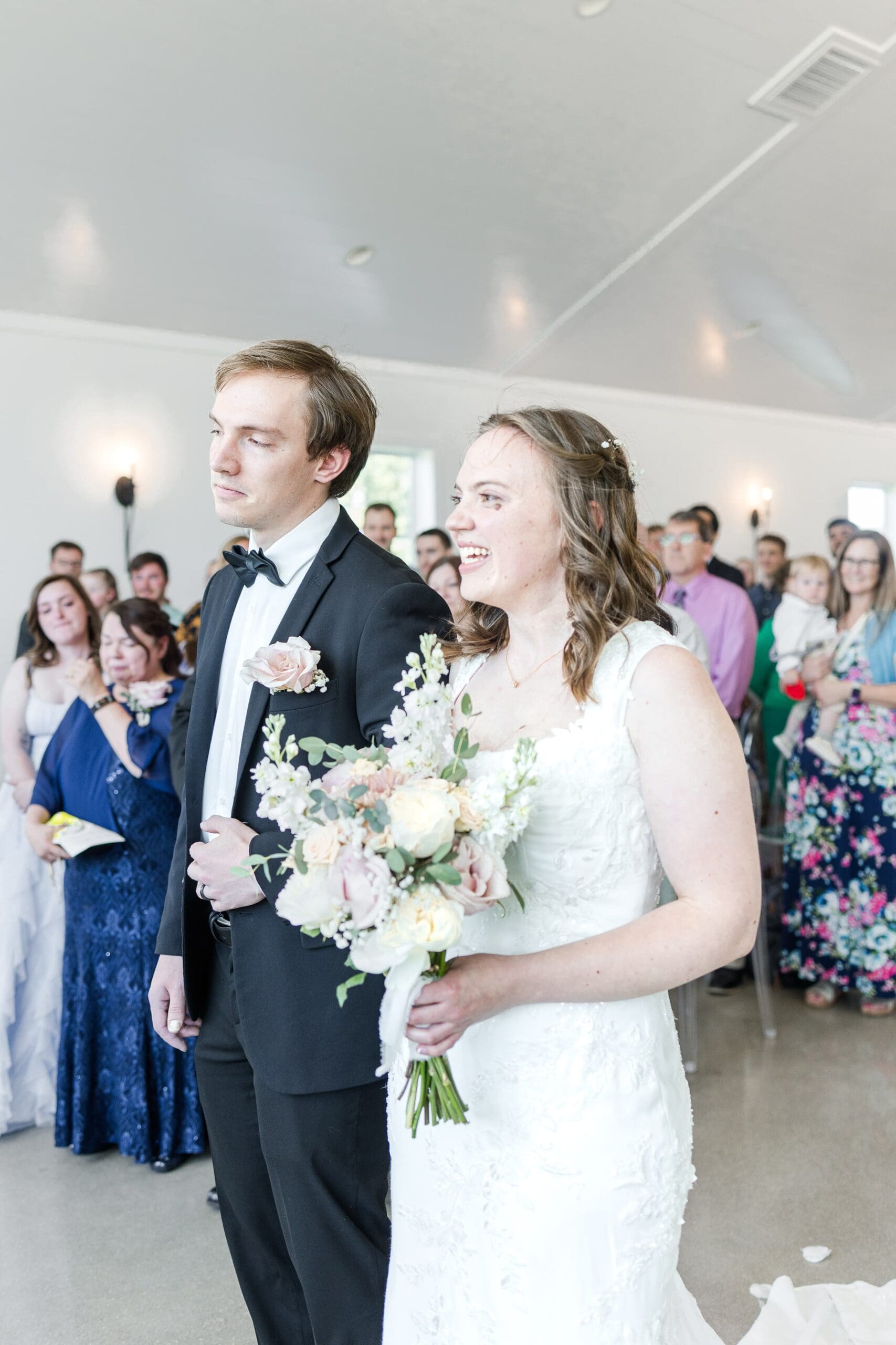 A bride in a white dress walks down the aisle of a bright, spacious hall filled with standing guests. People stand, smiling and capturing the moment.