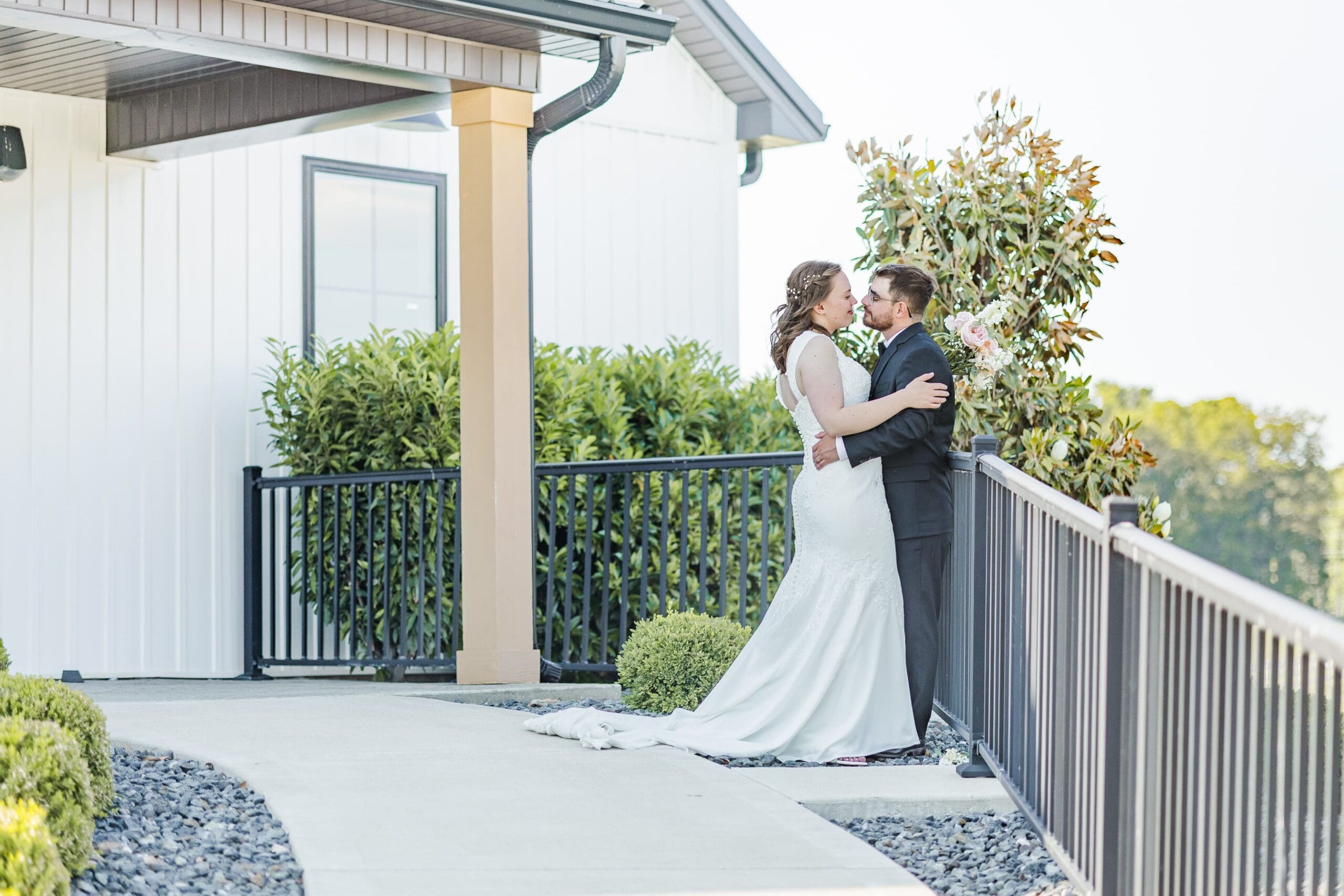 A newlywed couple embraces in front of a white building against a black railing and lush trees in the background. The groom and bride face each other and lean in close, almost kissing, both smiling warmly.