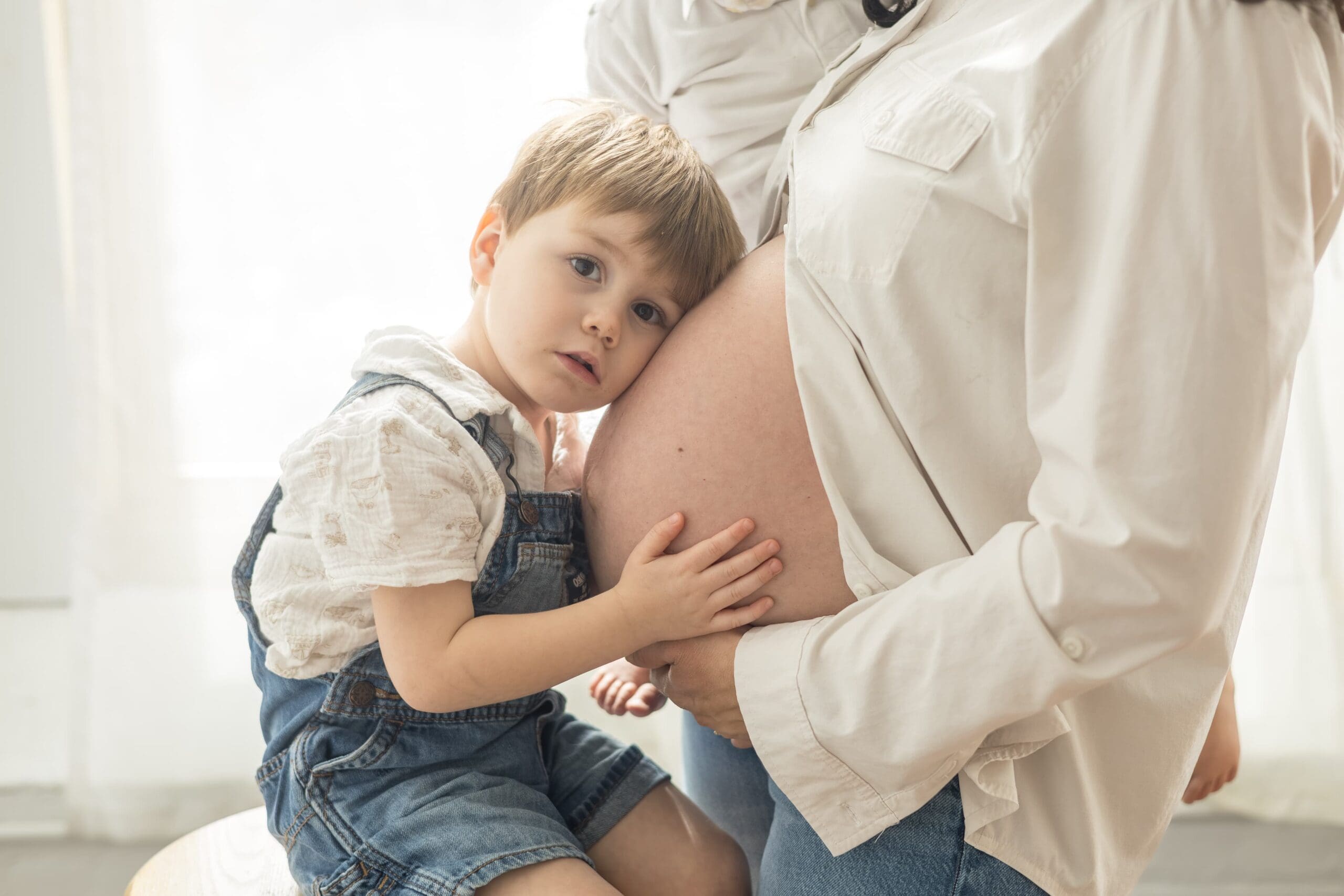 A young child in denim overalls gently leans on a pregnant person's belly, conveying warmth and tenderness. Both are wearing white shirts.