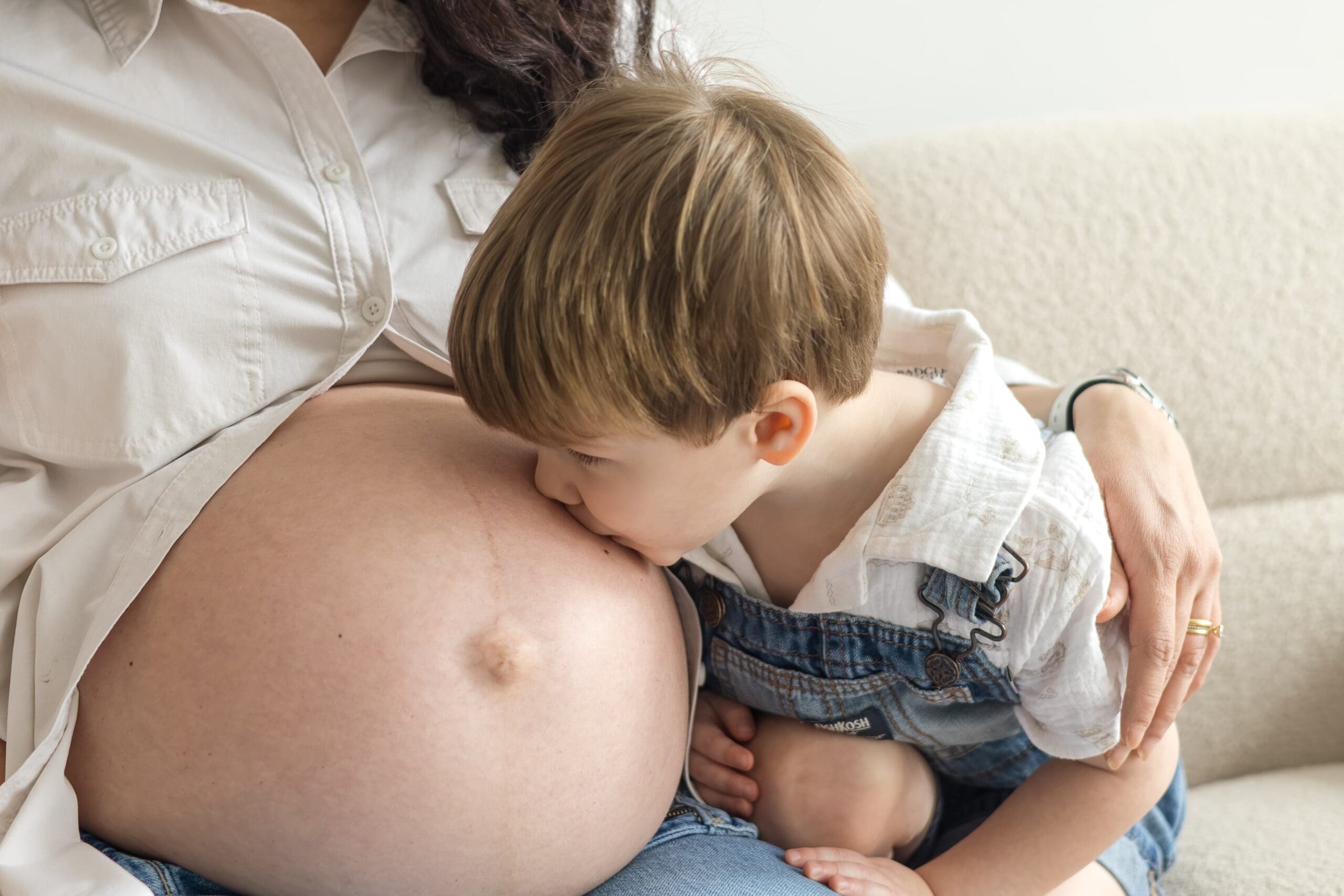A young child in denim overalls kisses on his mom's pregnant belly, conveying warmth and tenderness. Both are wearing white shirts.