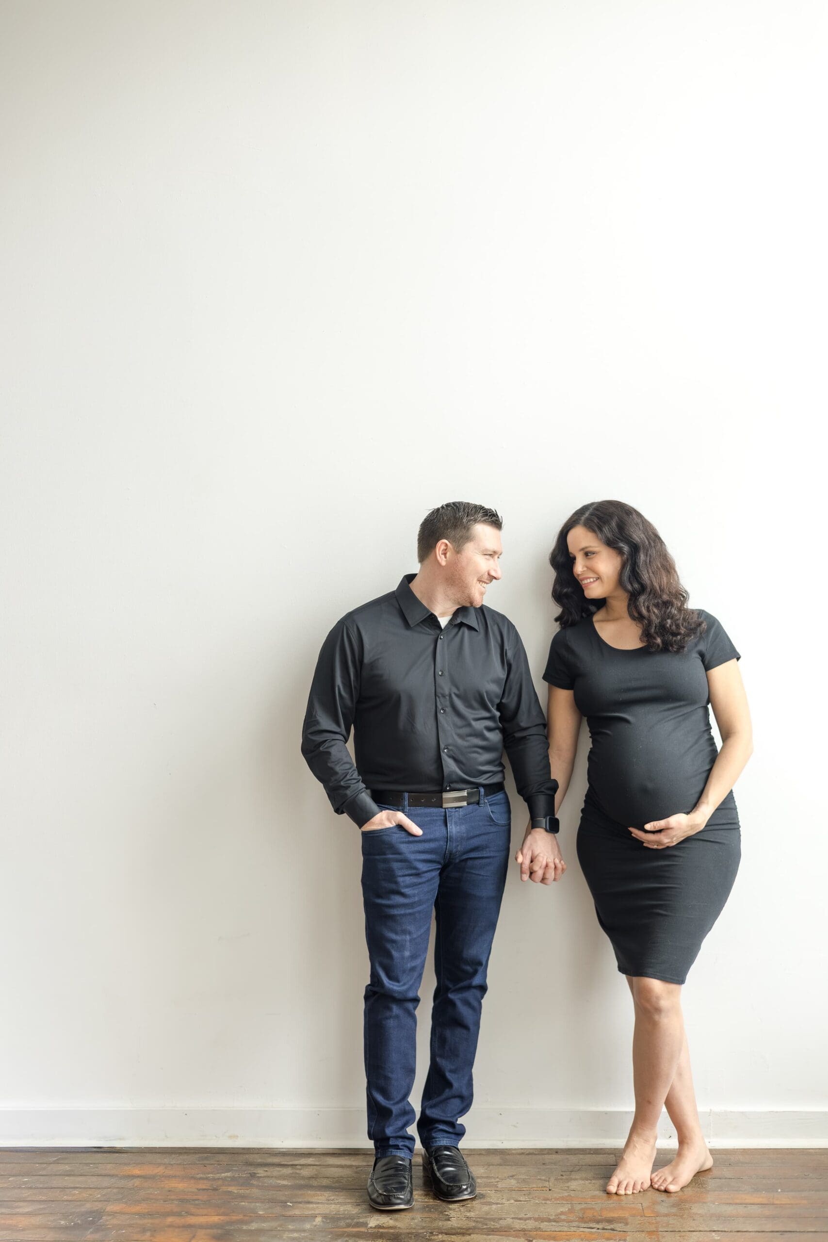 Husband and wife, both in black, hold hands and lean on a blank white wall while the woman holds her pregnant belly.