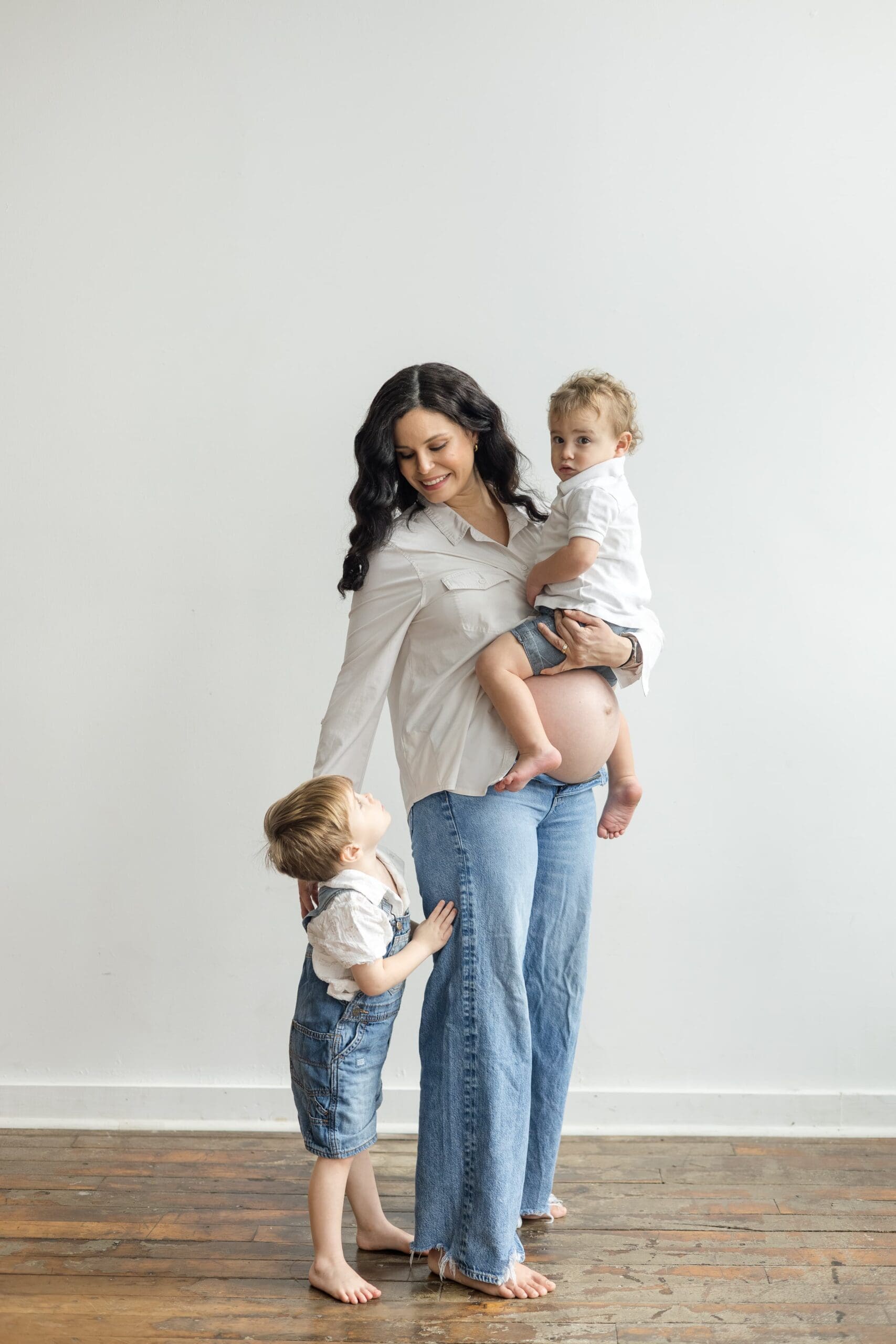 A smiling woman holds a toddler while another child stands beside her. They are in a bright, minimalistic room with a wooden floor, conveying warmth and family connection.