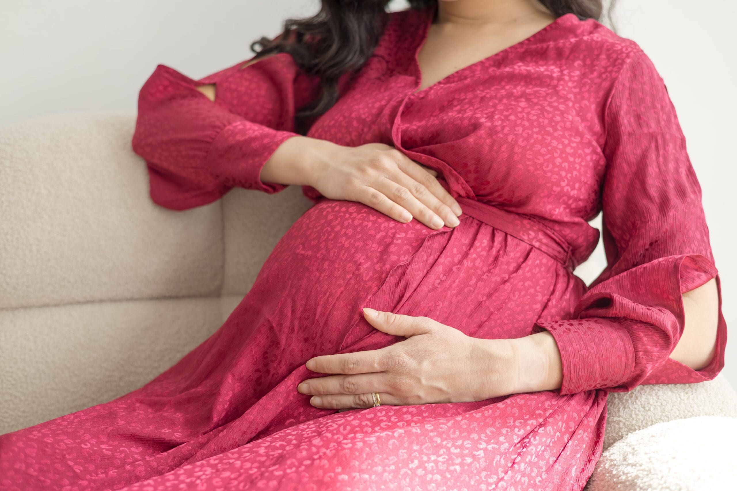 A pregnant woman in a flowing red dress cradles her pregnant belly on a tan couch.