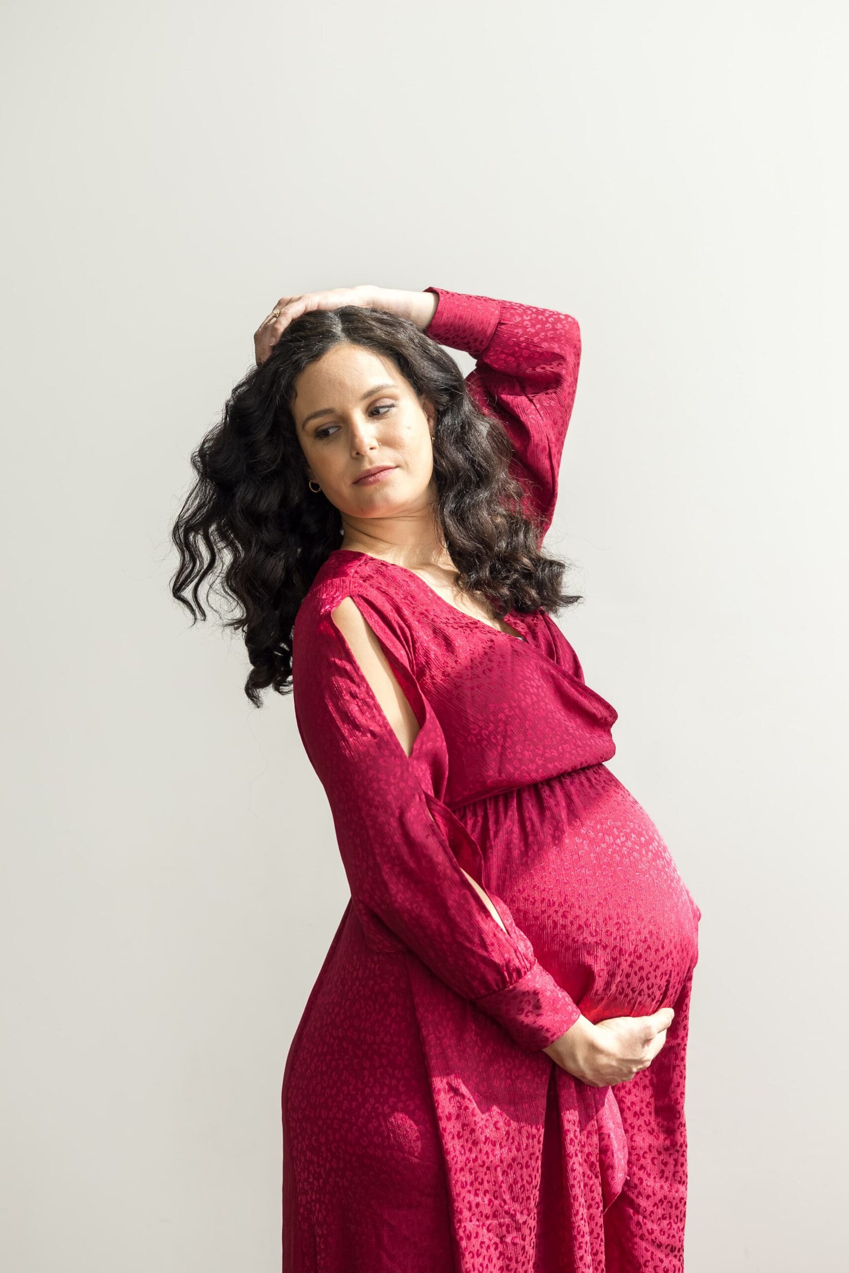 A pregnant woman in a flowing red dress gently cradles her belly with one hand and touches her curly hair with the other, exuding calm and confidence.