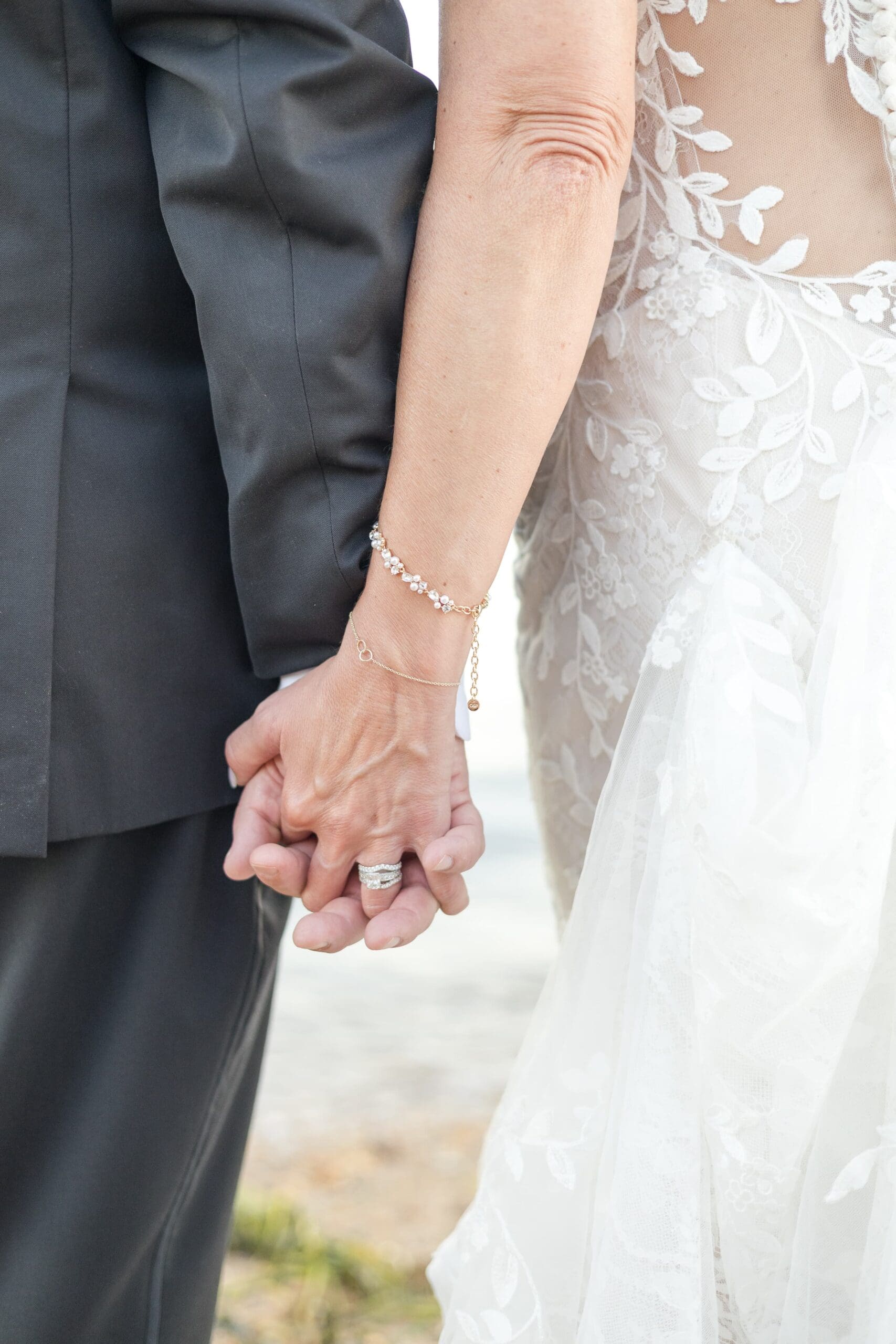 Close photo of a man and woman holding hands. They are in wedding attire, the man in a black suit and the woman in a lace white dress. Her ring displays as the main focus point.