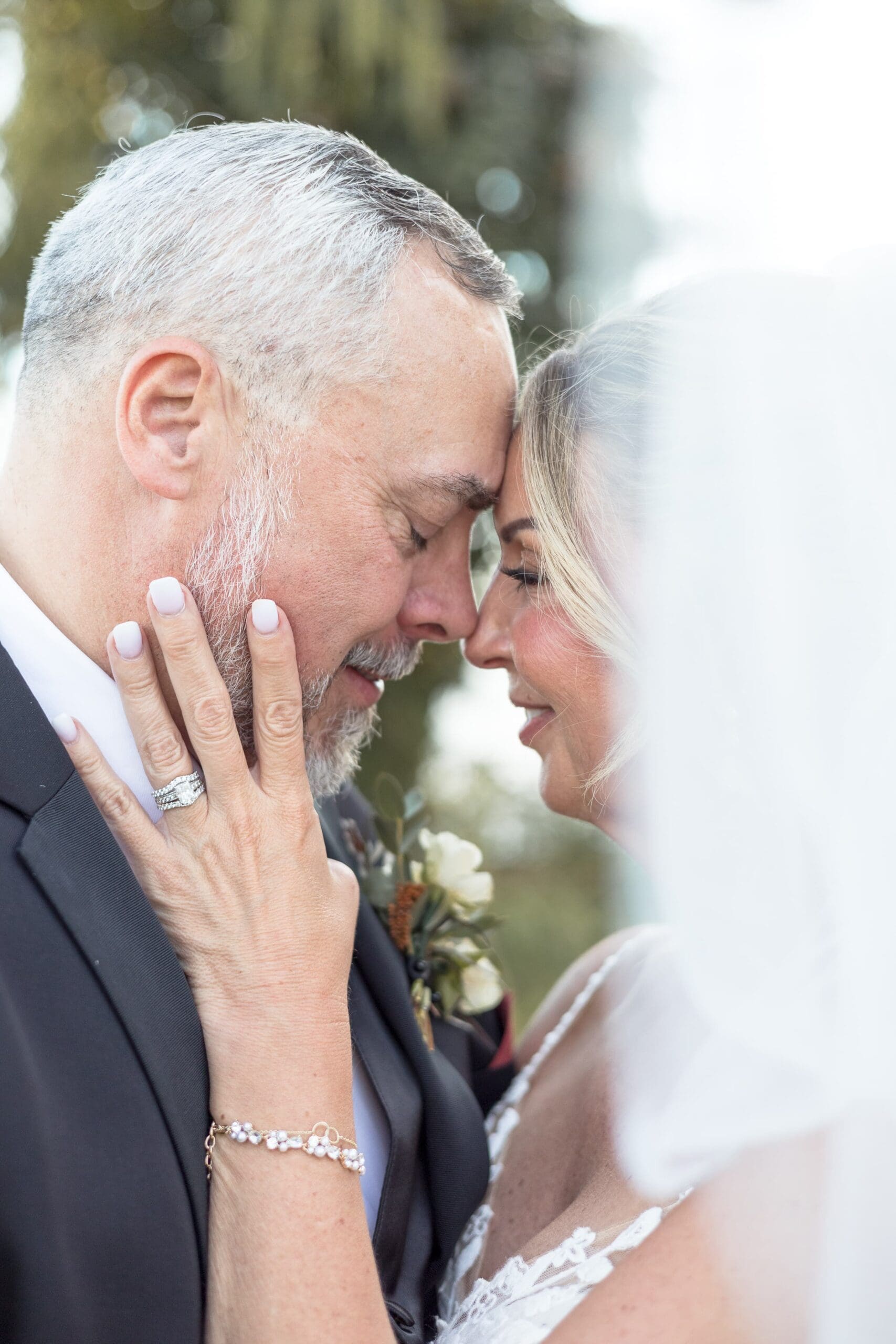 An older couple, in wedding attire, tenderly touch foreheads with eyes closed. The bride's hand, displaying a ring, rests on the groom's cheek, evoking deep affection.