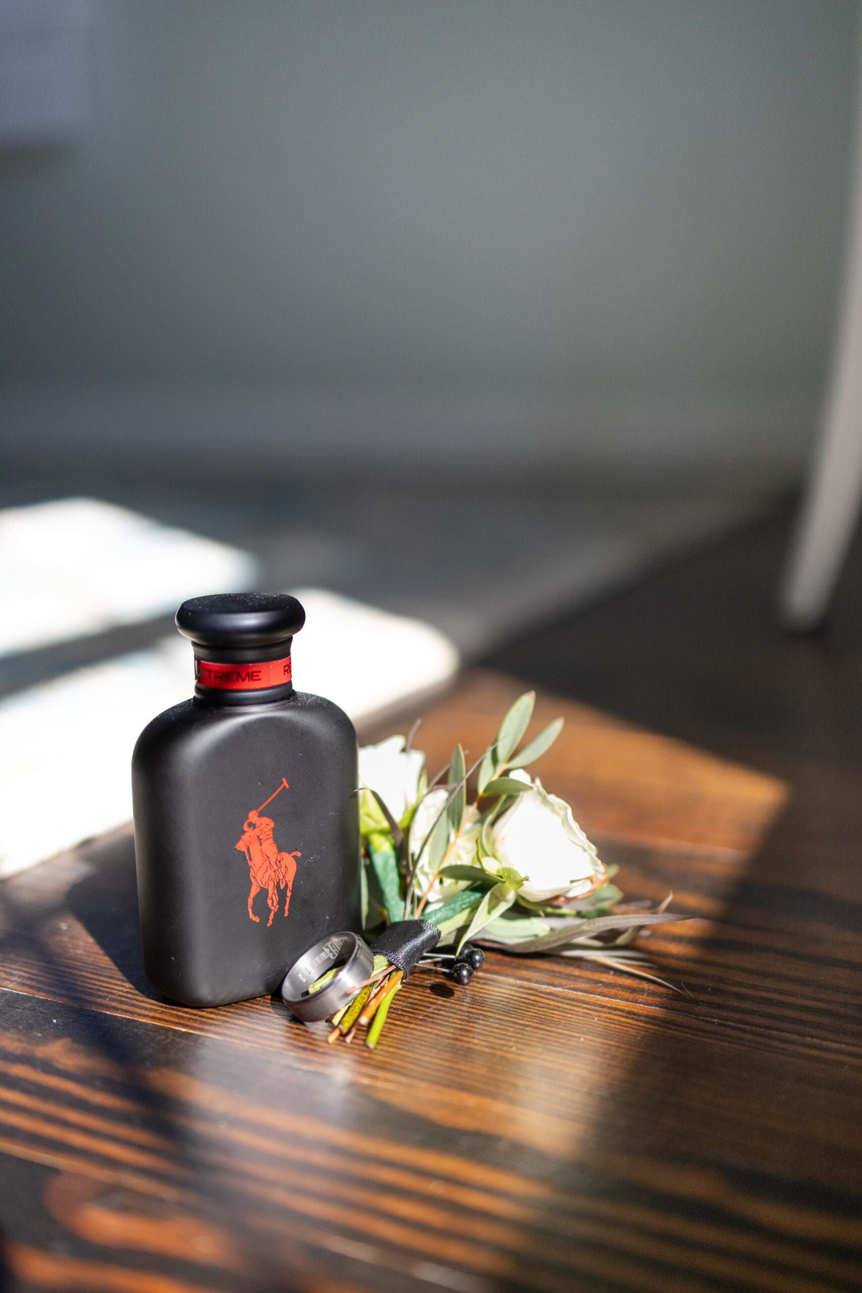 A cologne bottle, man's wedding ring and bouttonniere lay on a wooden ground, and the sun lights up the array.