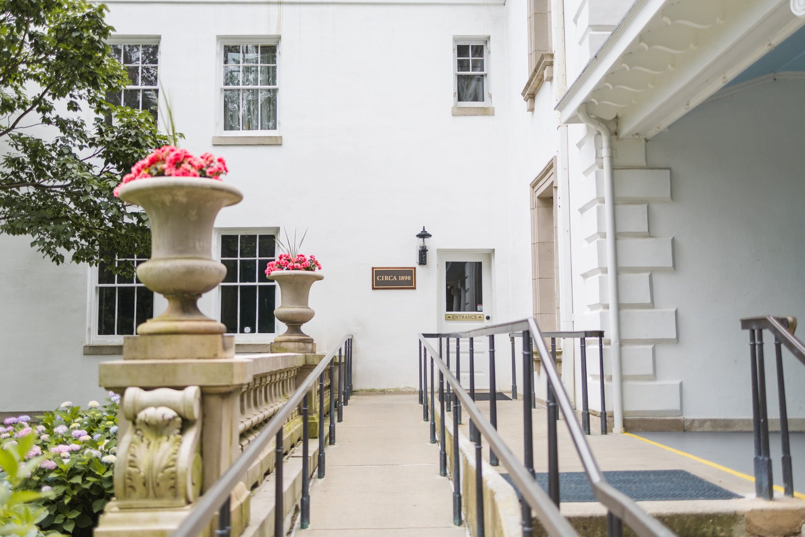 Wheelchair-accessible ramp leads to a white building entrance. Large planters with red flowers line the path. The scene is bright and welcoming.