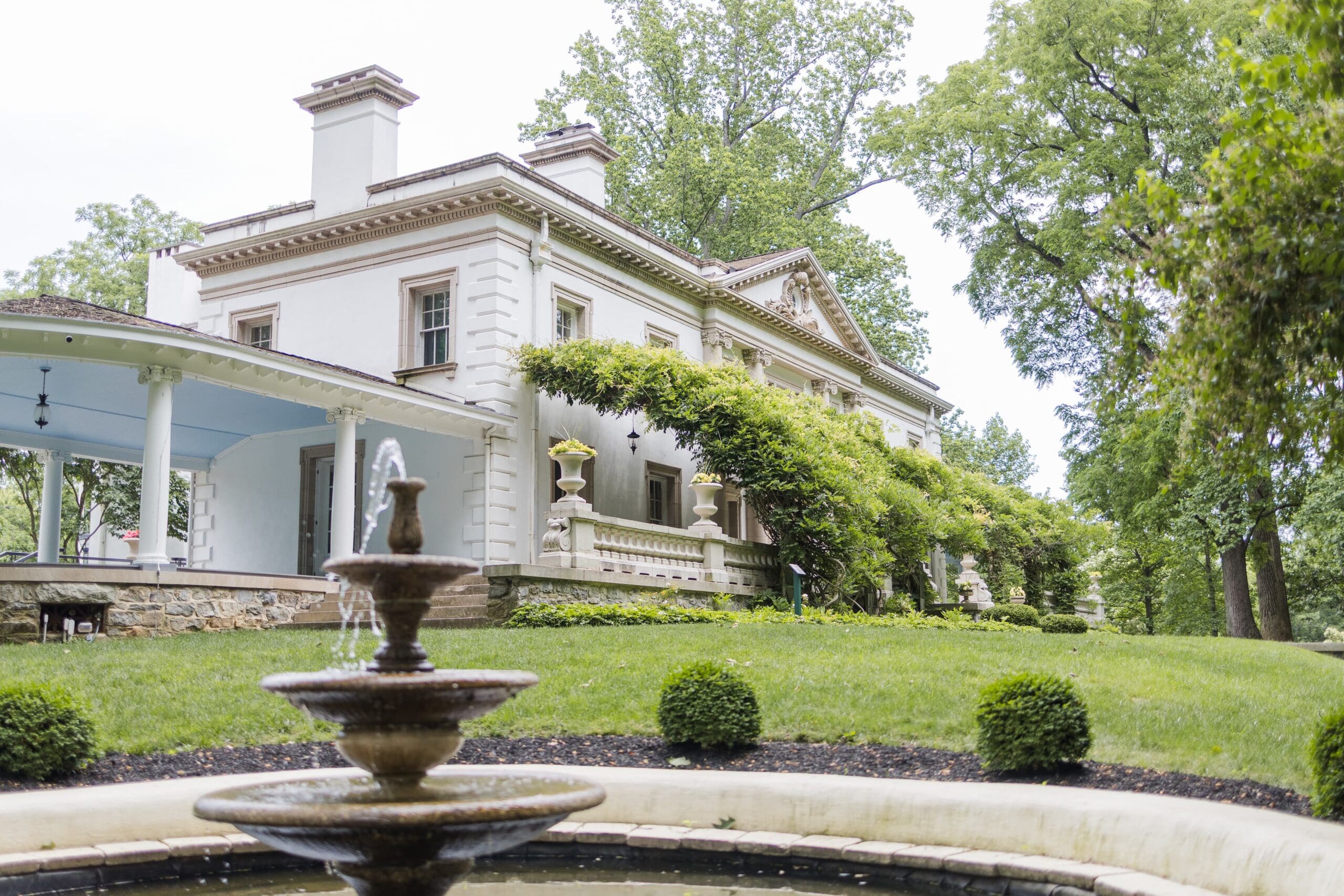 Elegant white mansion with wisteria-covered walls, large porch, and three-tiered fountain in the foreground. Surrounded by lush greenery and manicured bushes.
