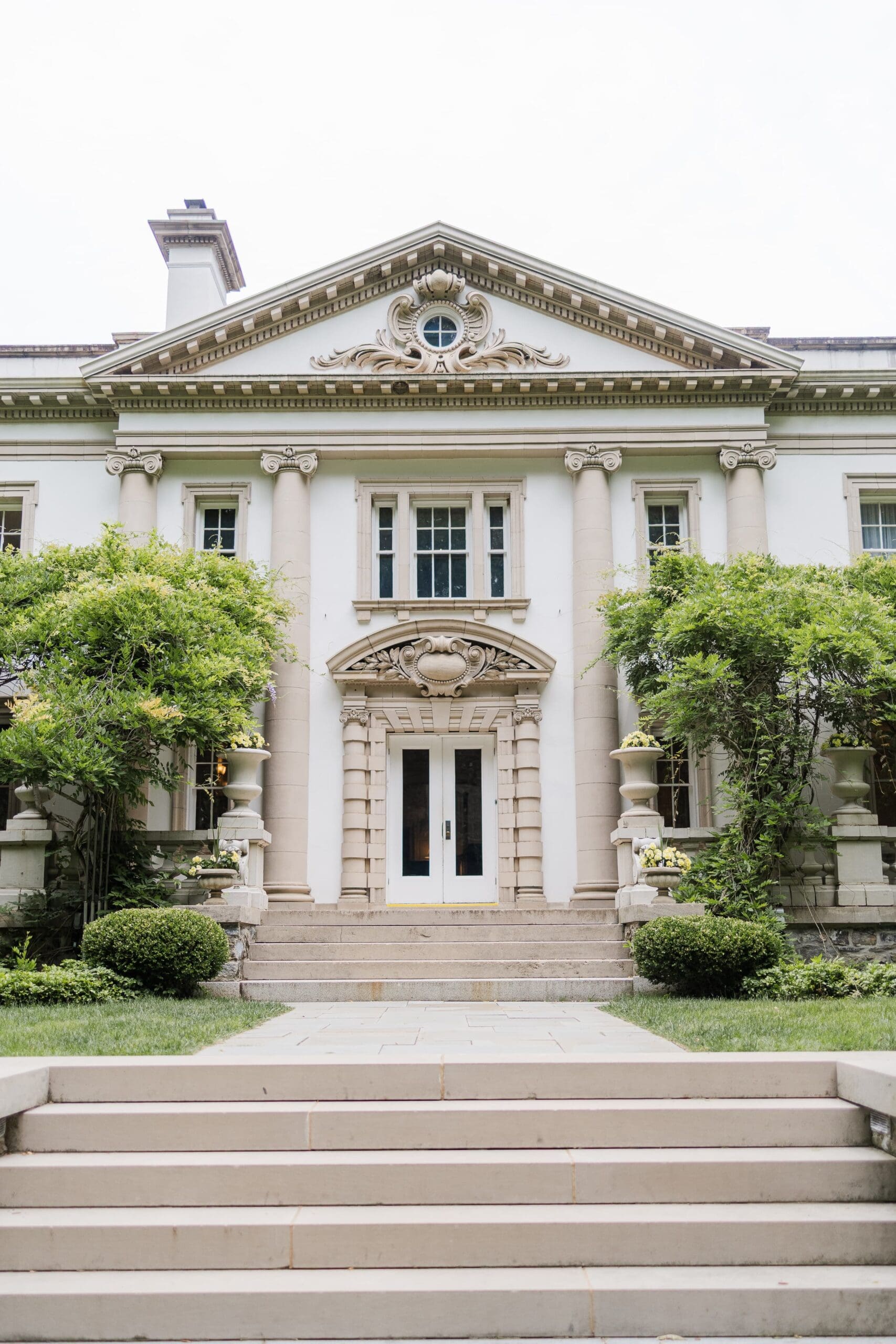Elegant white mansion with wisteria-covered walls, large porch. Surrounded by lush greenery and manicured bushes.