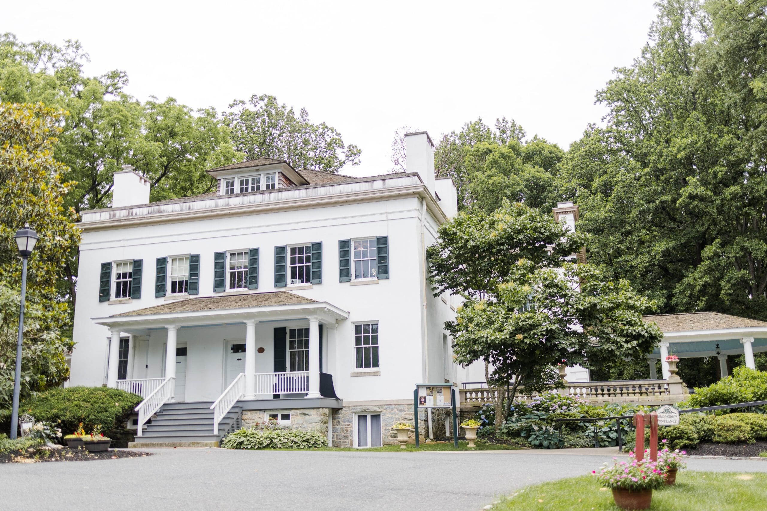 Liriodendron, an Elegant white mansion with a large covered porch. Surrounded by lush greenery and manicured bushes.