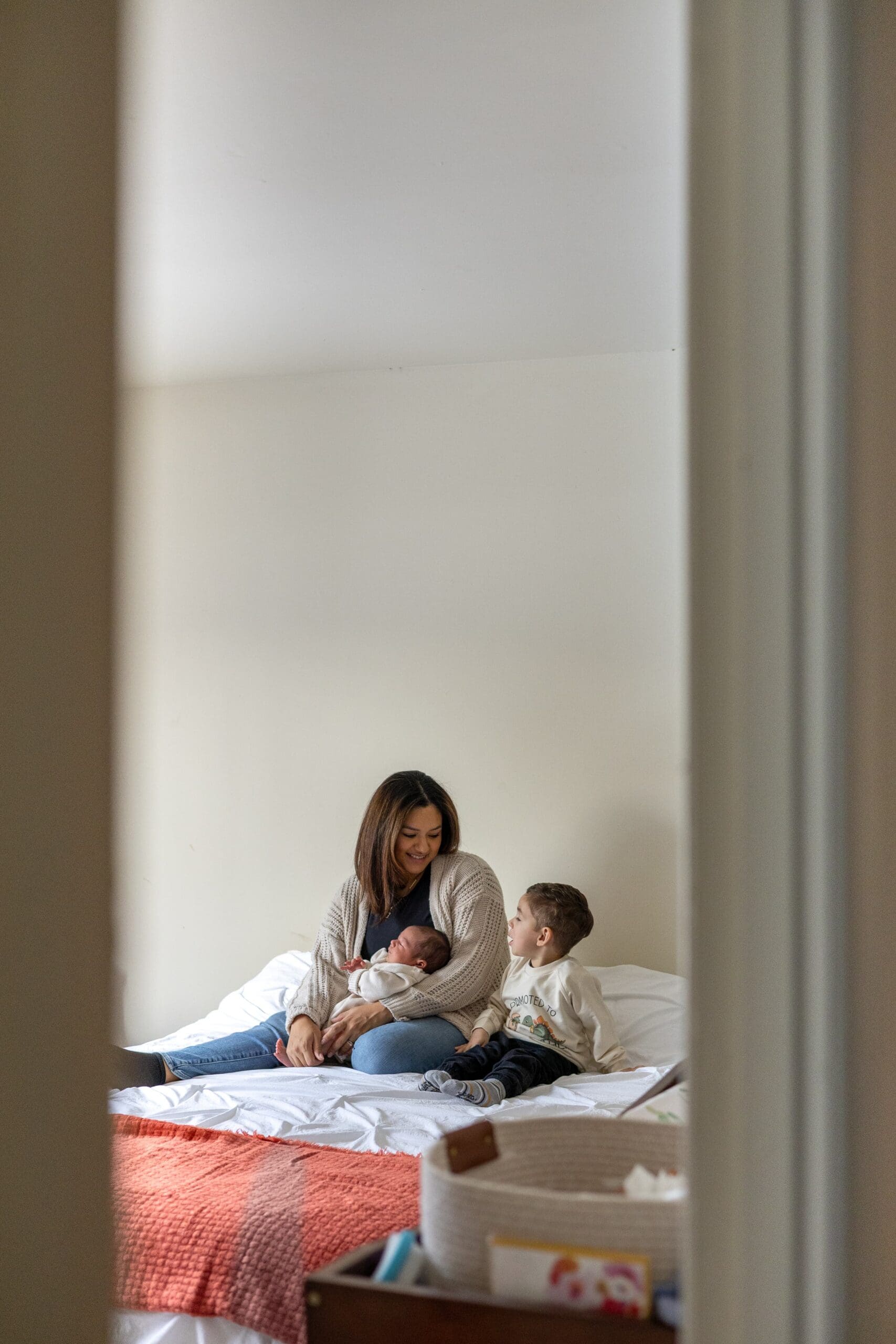 A woman sits on a white bed, smiling at her son while she holds a baby. The young child also on the bed, smiles up at her, both wearing cozy, neutral-toned sweaters.