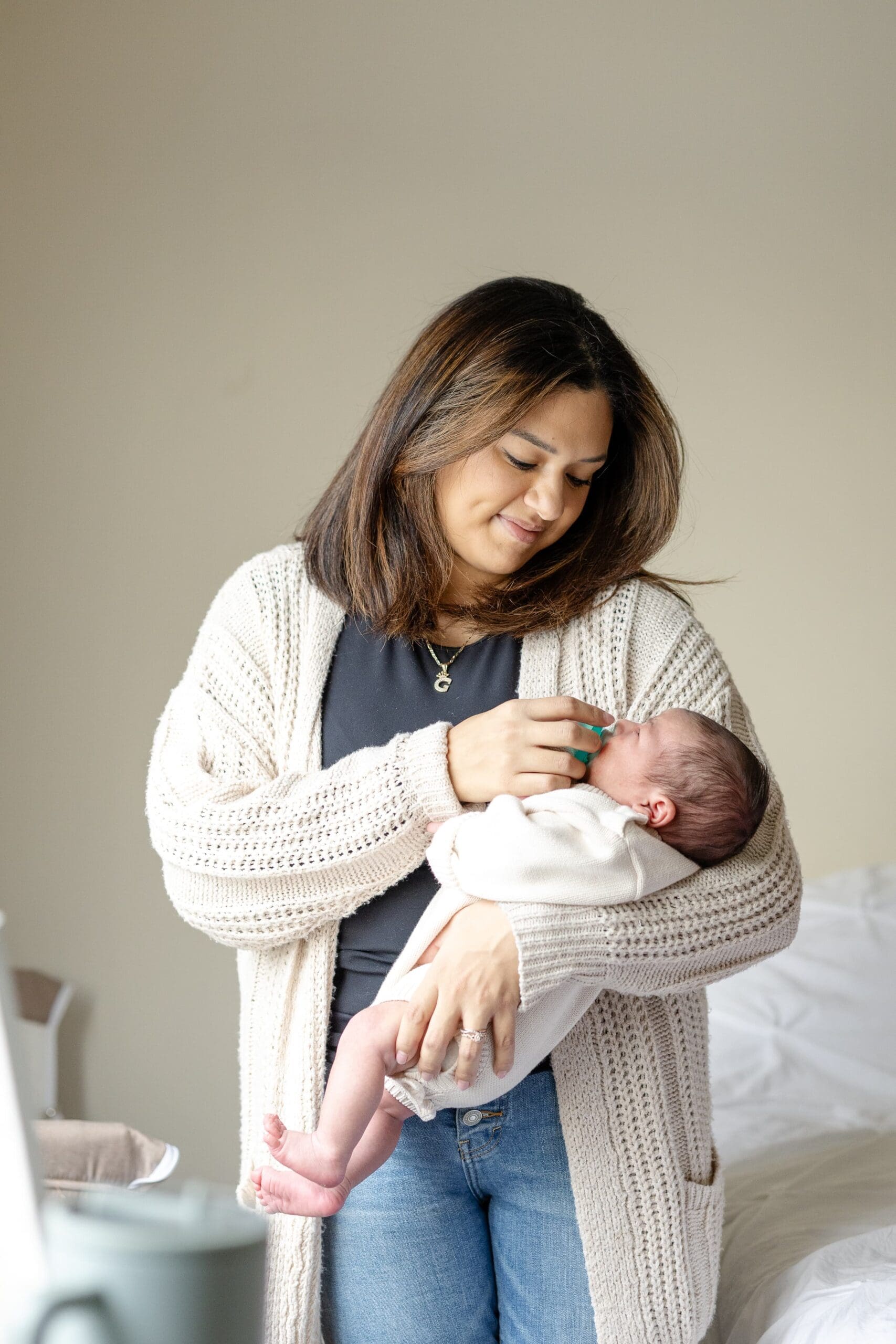A woman gently holds a sleeping baby in her arms, offering a pacifier. She wears a cozy sweater, standing in a softly lit room with a warm, caring expression.