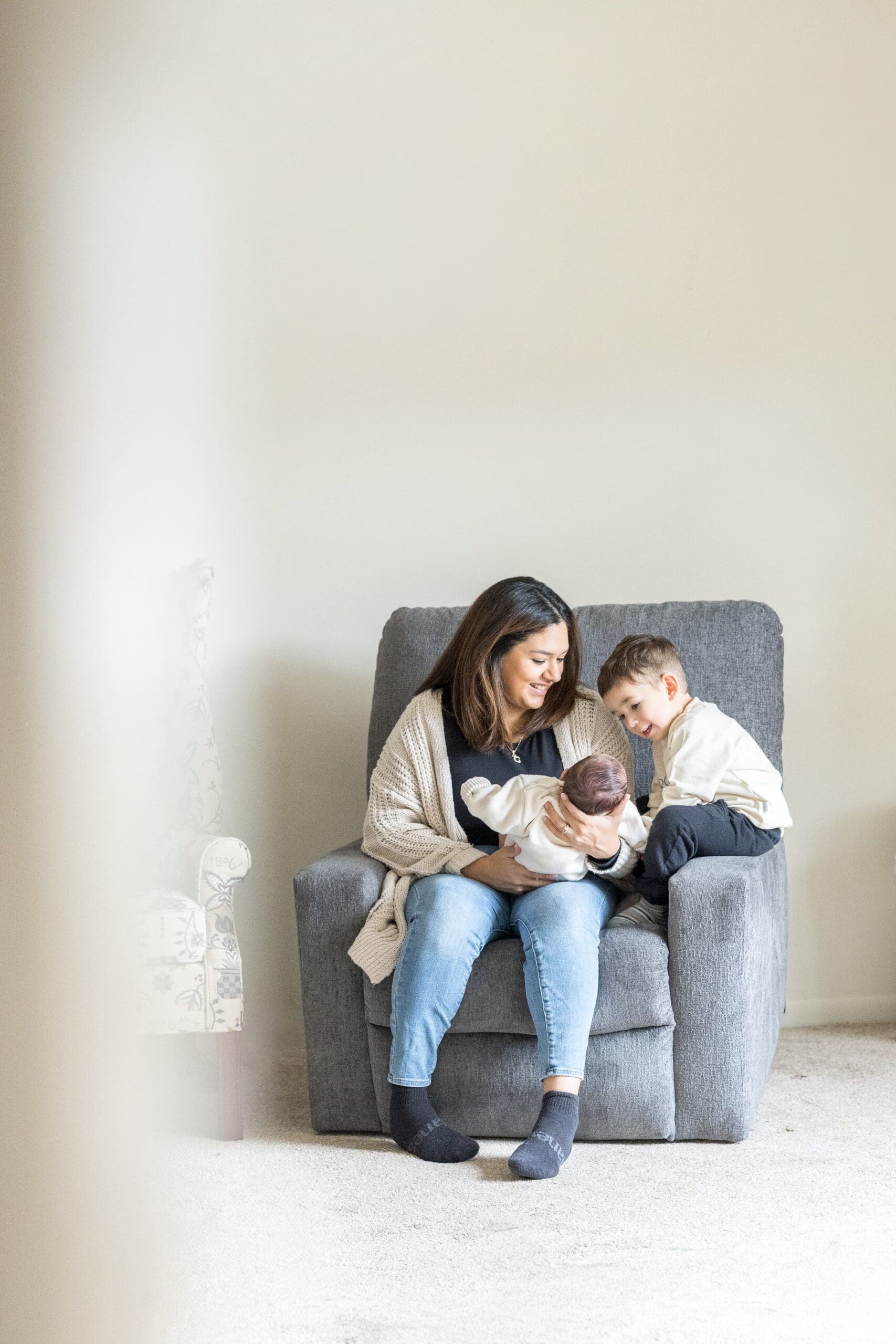 A woman sits on a gray armchair, smiling at a baby she's holding. A young child leans in, watching, both wearing cozy, neutral-toned sweaters.