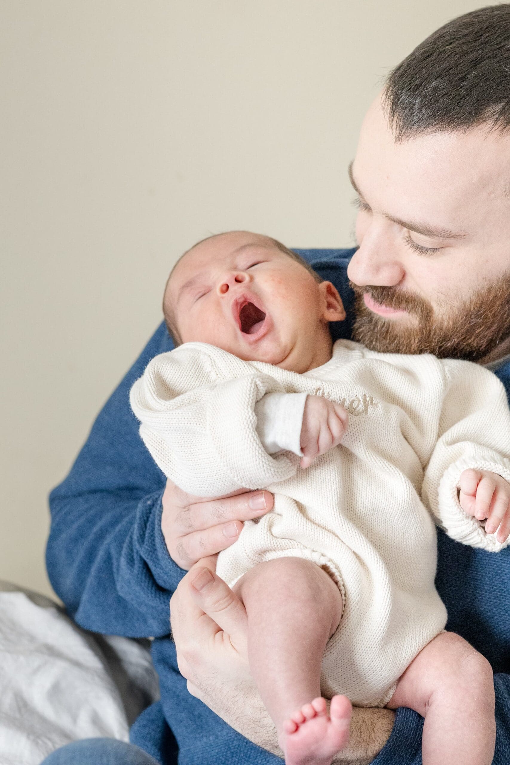 A bearded man gently holds a yawning baby in his arms, smiling at him. He wears a cozy sweater, sitting in a softly lit room with a warm, caring expression.