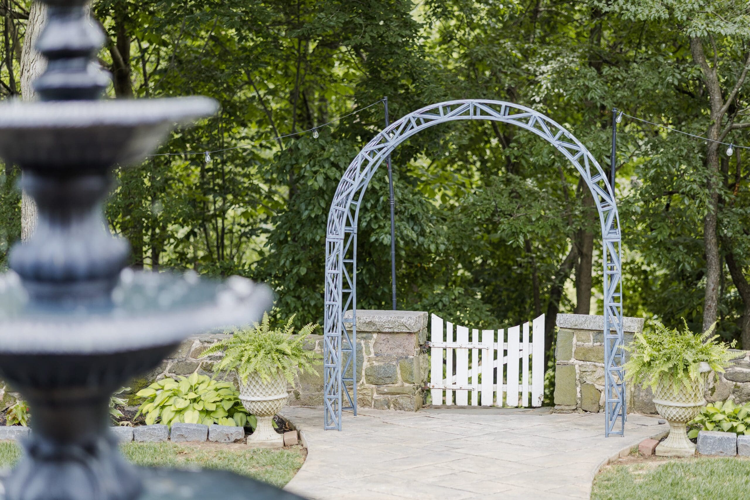 A garden scene with an elegant metal archway and a white picket gate framed by lush green trees. A blurred fountain is in the foreground, adding depth.