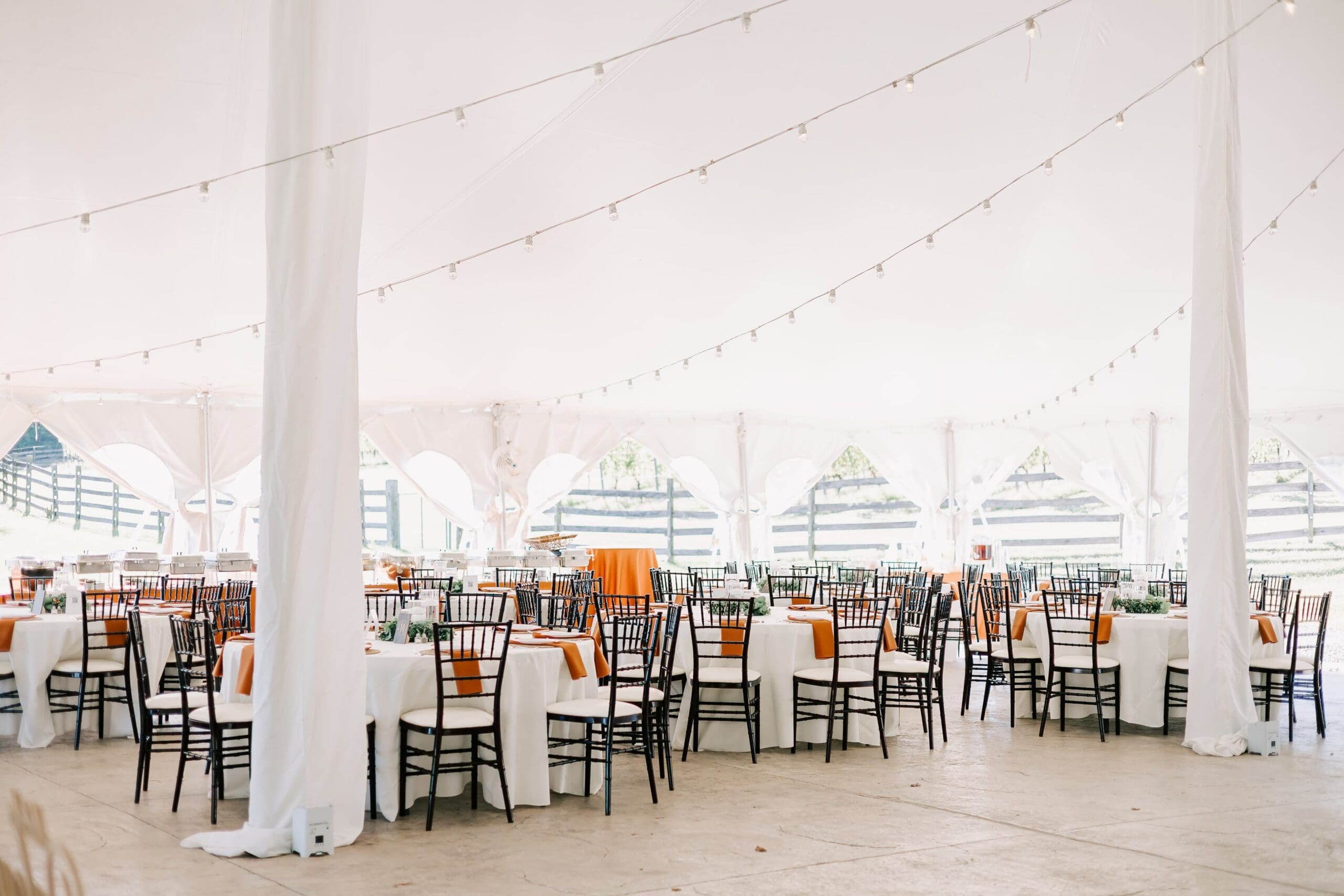 Elegant wedding reception under a white tent with round tables, black chairs, white linens, and string lights overhead, creating a festive atmosphere.