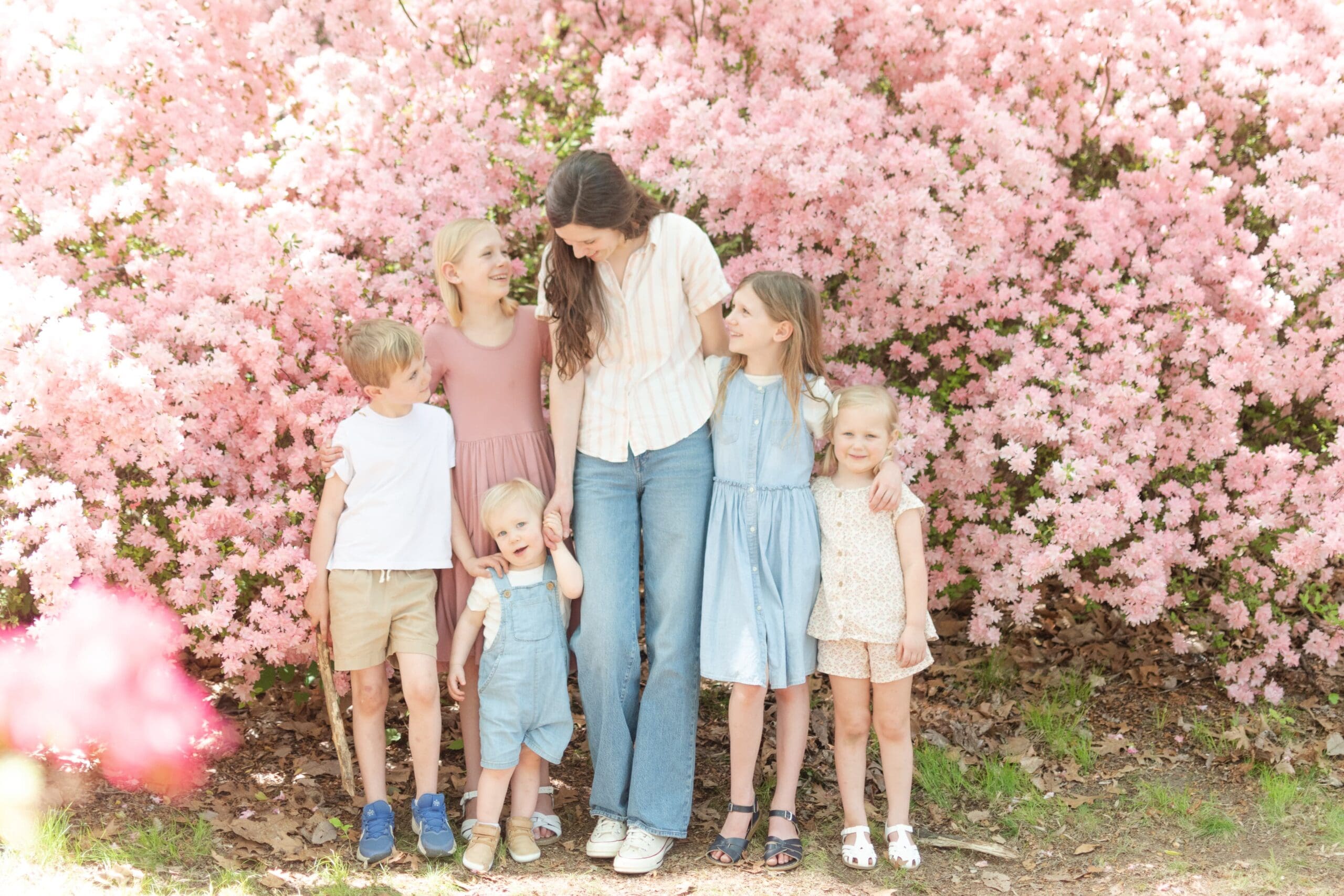 A woman and five children stand closely together, smiling in front of vibrant pink flowers. The scene conveys warmth and family joy amidst nature.