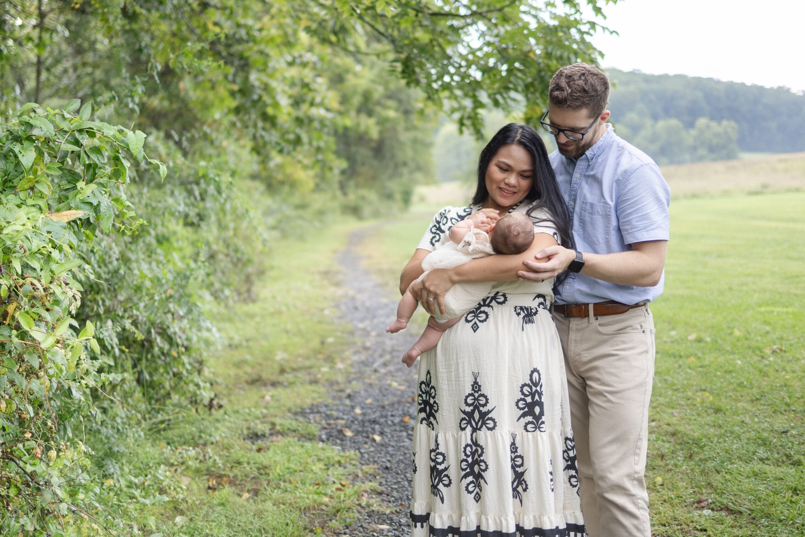 A couple stands on a grassy path, lovingly holding a baby. They are surrounded by lush greenery, conveying warmth and family togetherness.