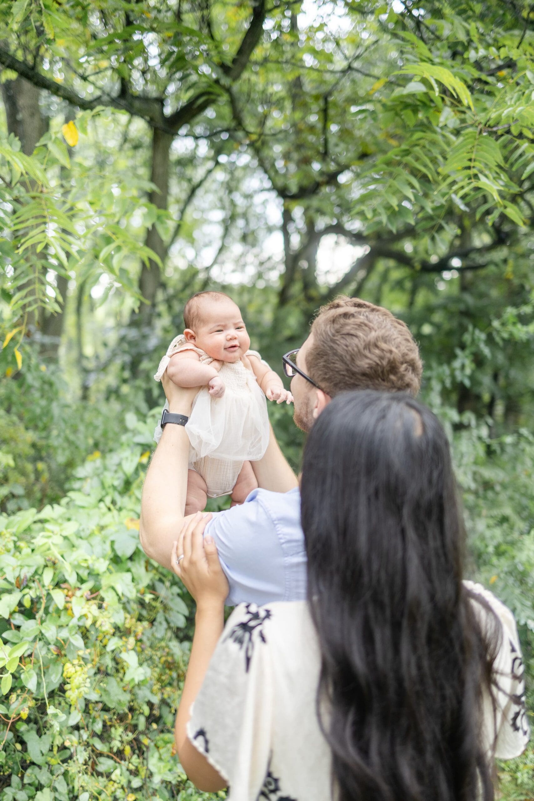 A couple stands on a grassy path, lovingly holding a baby in the air. They are surrounded by lush greenery, conveying warmth and family togetherness.