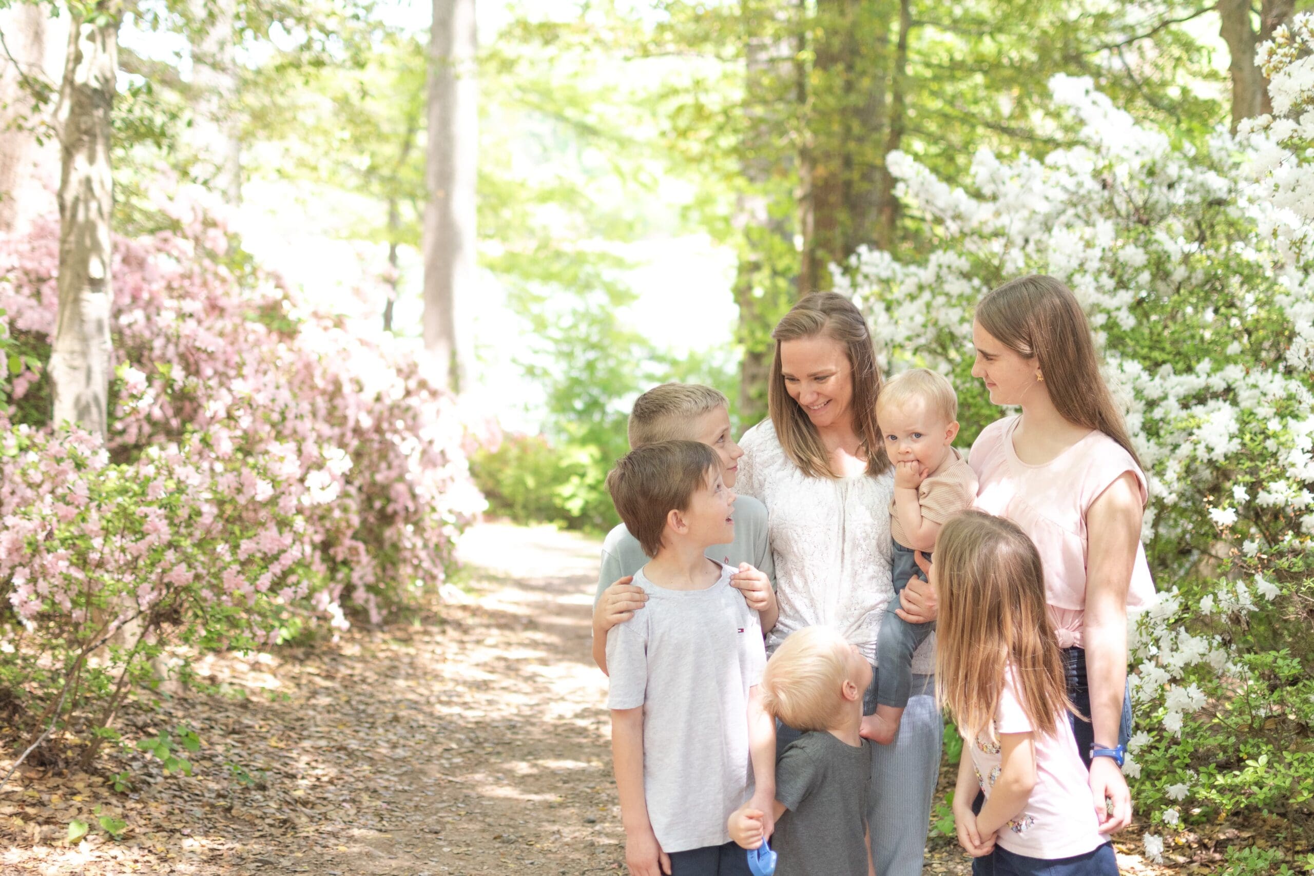 A woman and five children stand closely together, smiling in front of vibrant pink flowers. The scene conveys warmth and family joy amidst nature.