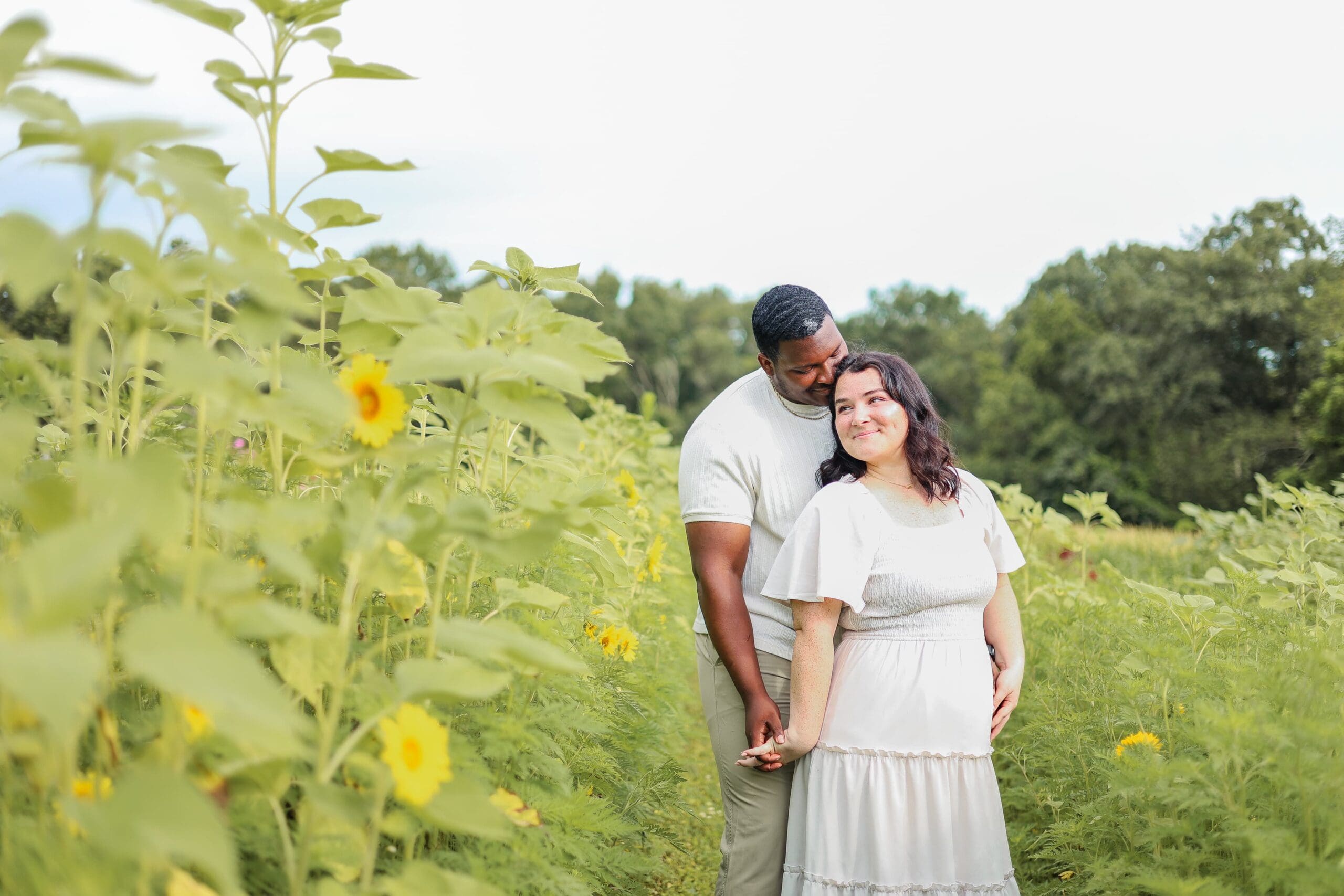 A couple stands in a sunlit meadow of sunflowers, embracing and smiling. The landscape is lush and green, conveying a warm and joyful atmosphere.