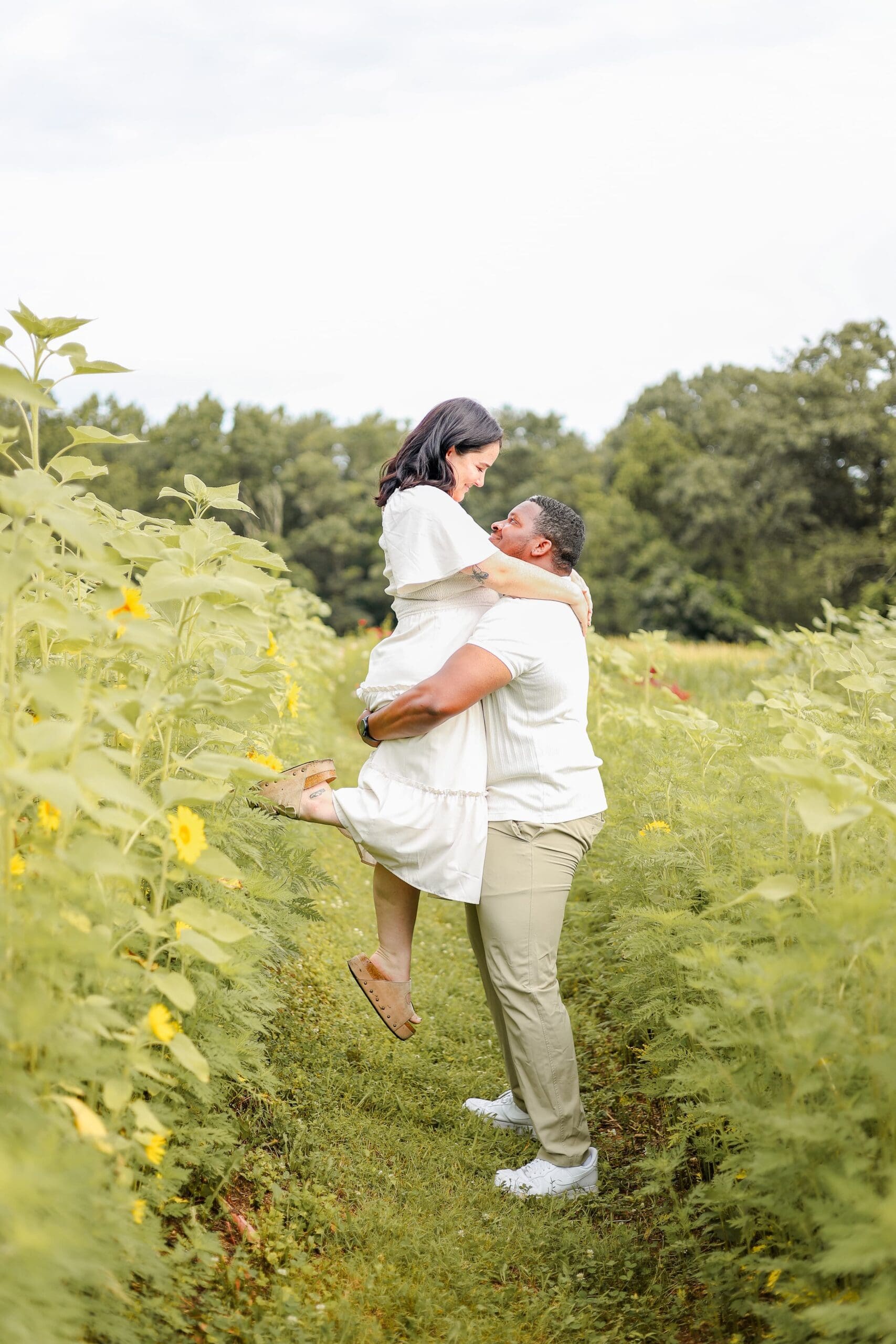 A couple stands in a sunlit meadow of sunflowers, embracing and smiling. The man lifts her up as she pops her heel. The landscape is lush and green, conveying a warm and joyful atmosphere.