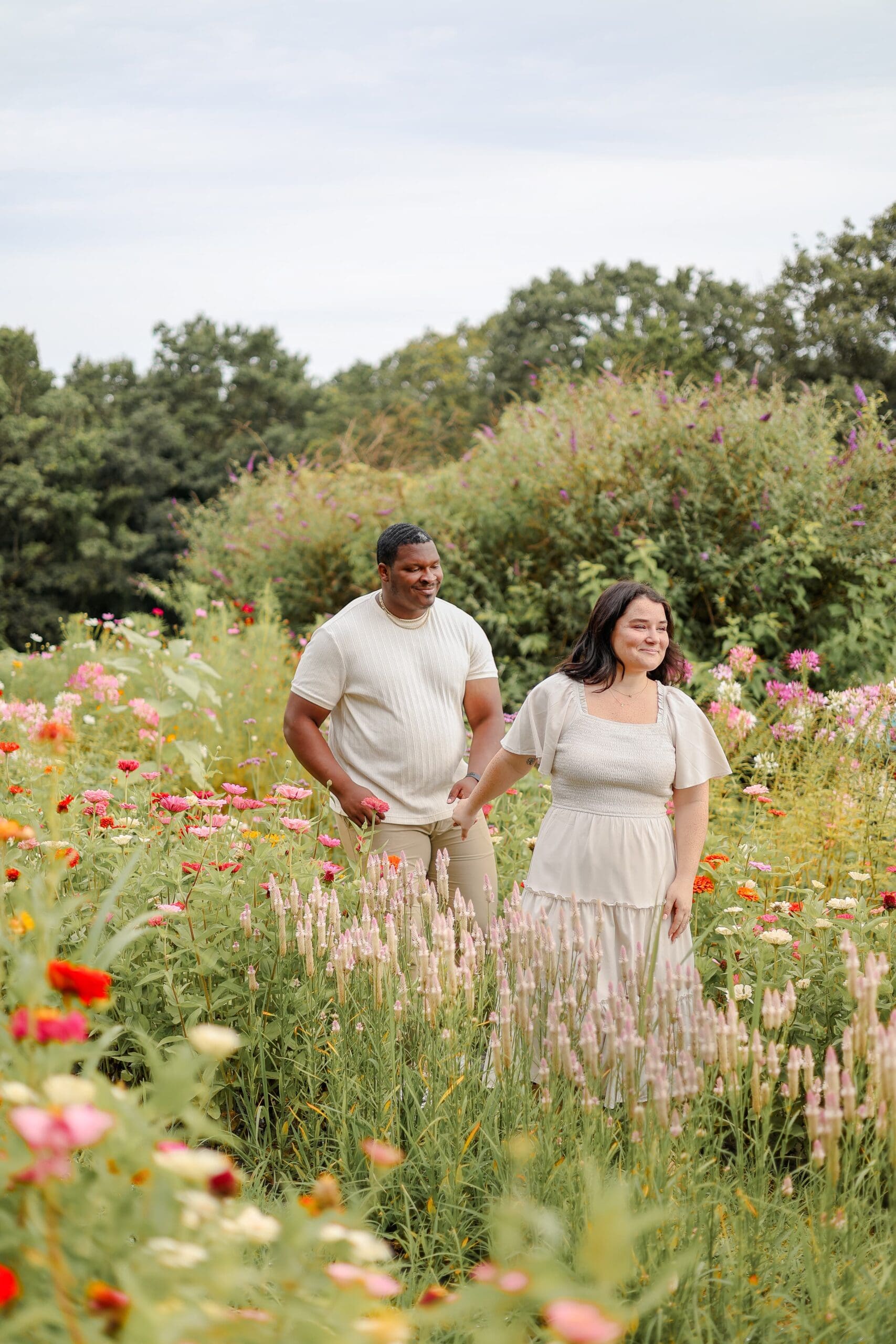 A couple walks and smiles out in a vibrant flower field. They wear light, casual clothing. The scene feels joyful and tranquil.