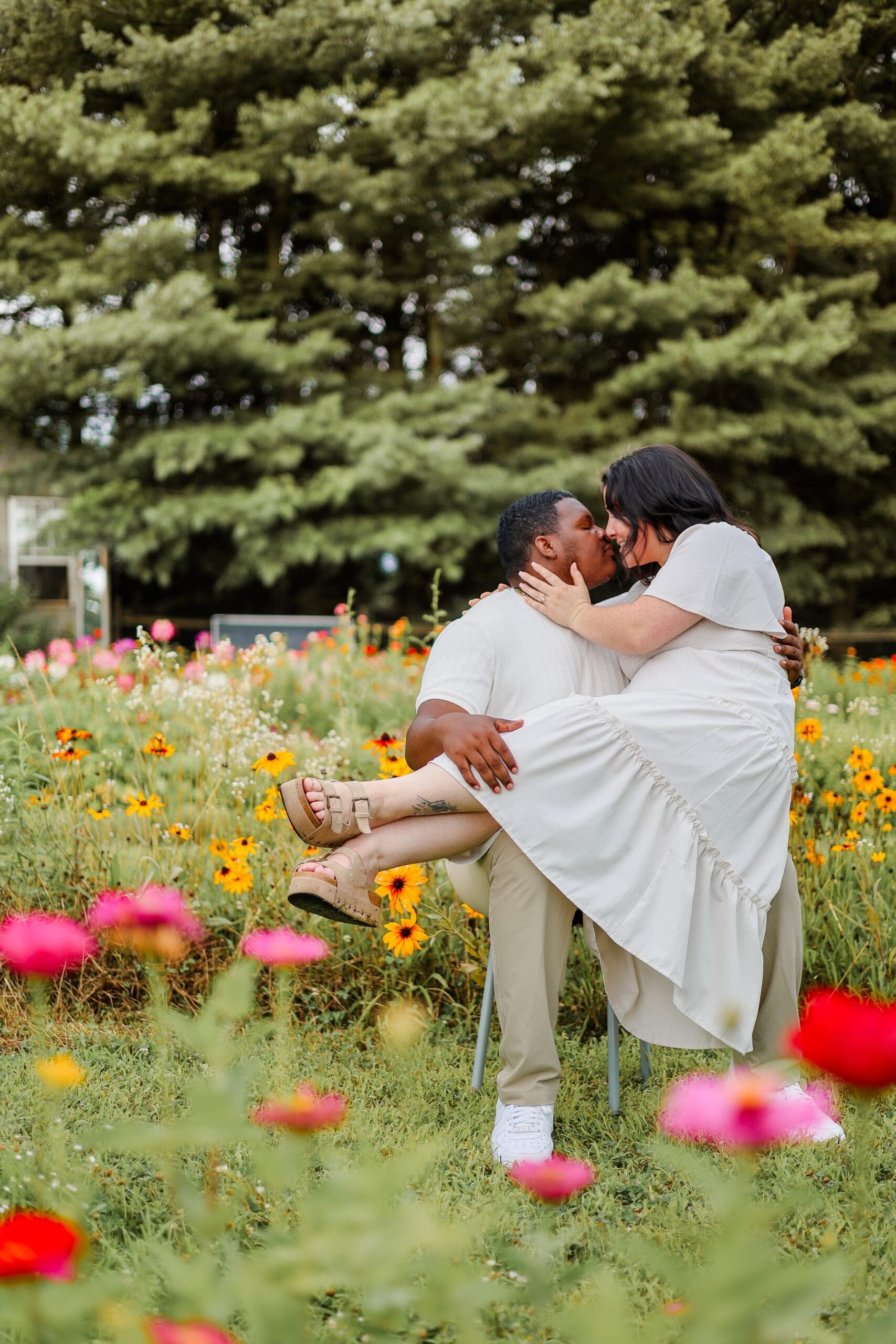 A couple sits and smiles at each other in a vibrant flower field. The woman has her hand on the man's cheek. They wear light, casual clothing. The scene feels joyful and tranquil.