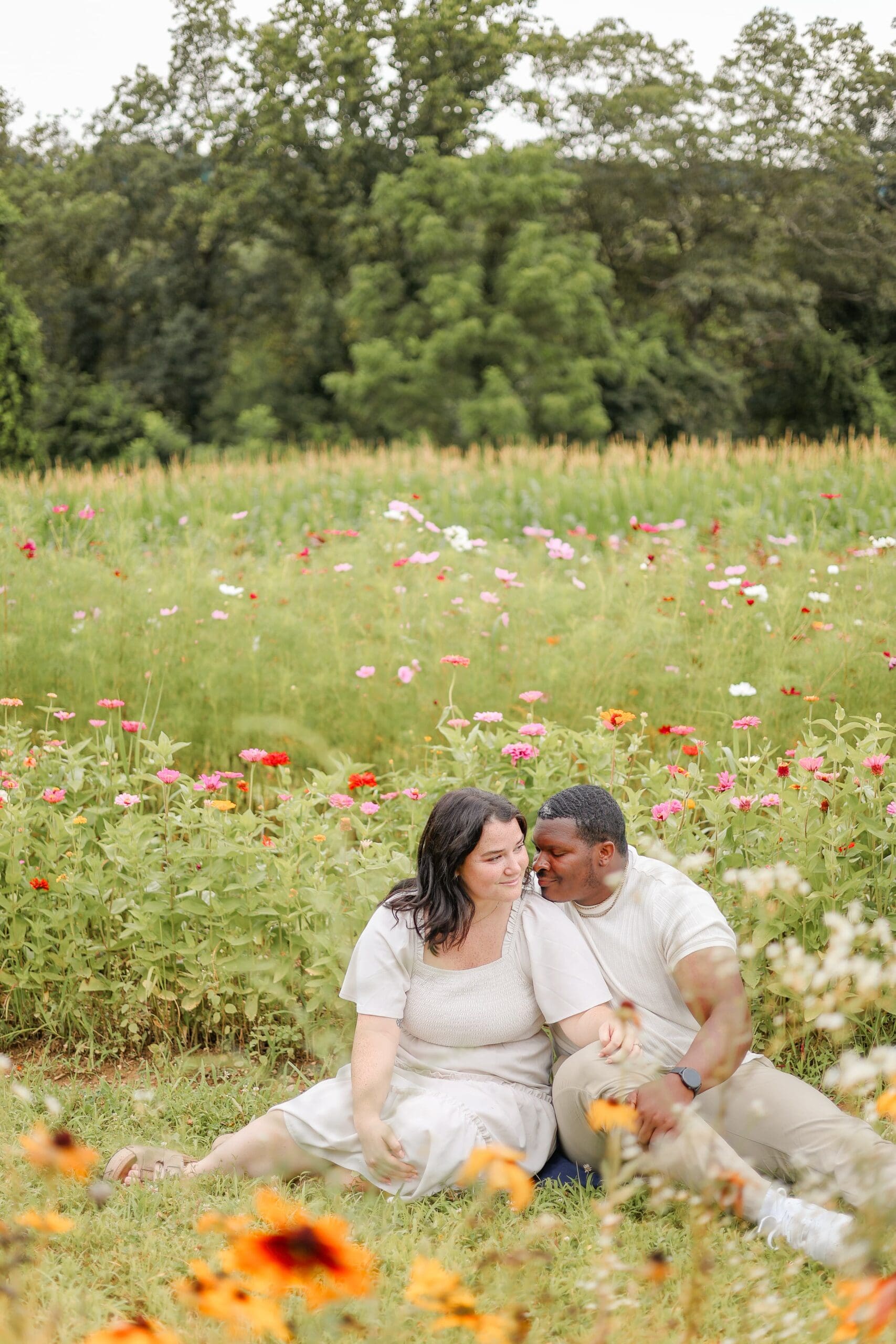 A couple sits close and smiles at each other in a vibrant flower field. They wear light, casual clothing. The scene feels joyful and tranquil.