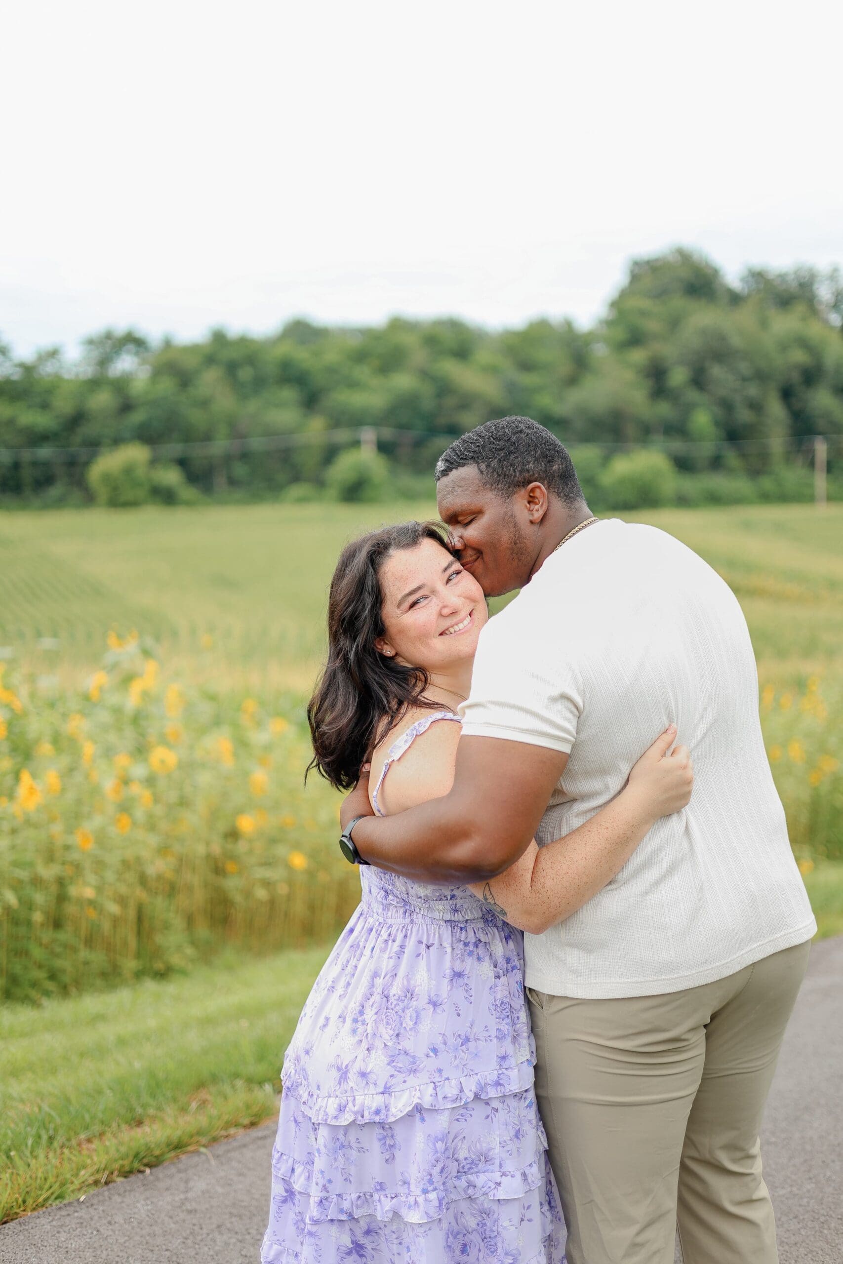 A couple stands in a sunlit meadow of sunflowers, embracing and smiling. The fiance kisses her cheek. The landscape is lush and green, conveying a warm and joyful atmosphere.