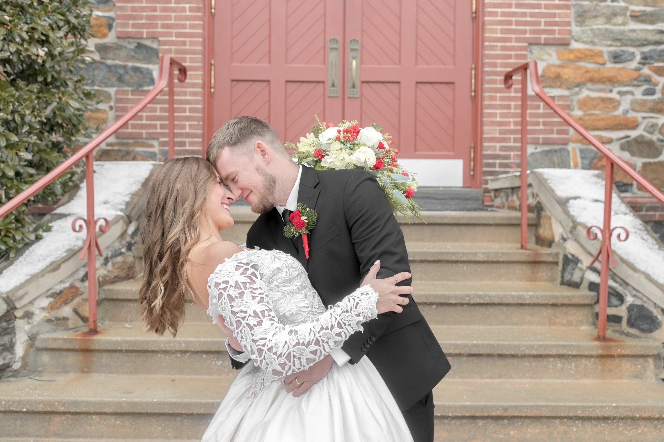Woman holding a floral bouquet over her husbands neck, posed in front of red doors and brick church.