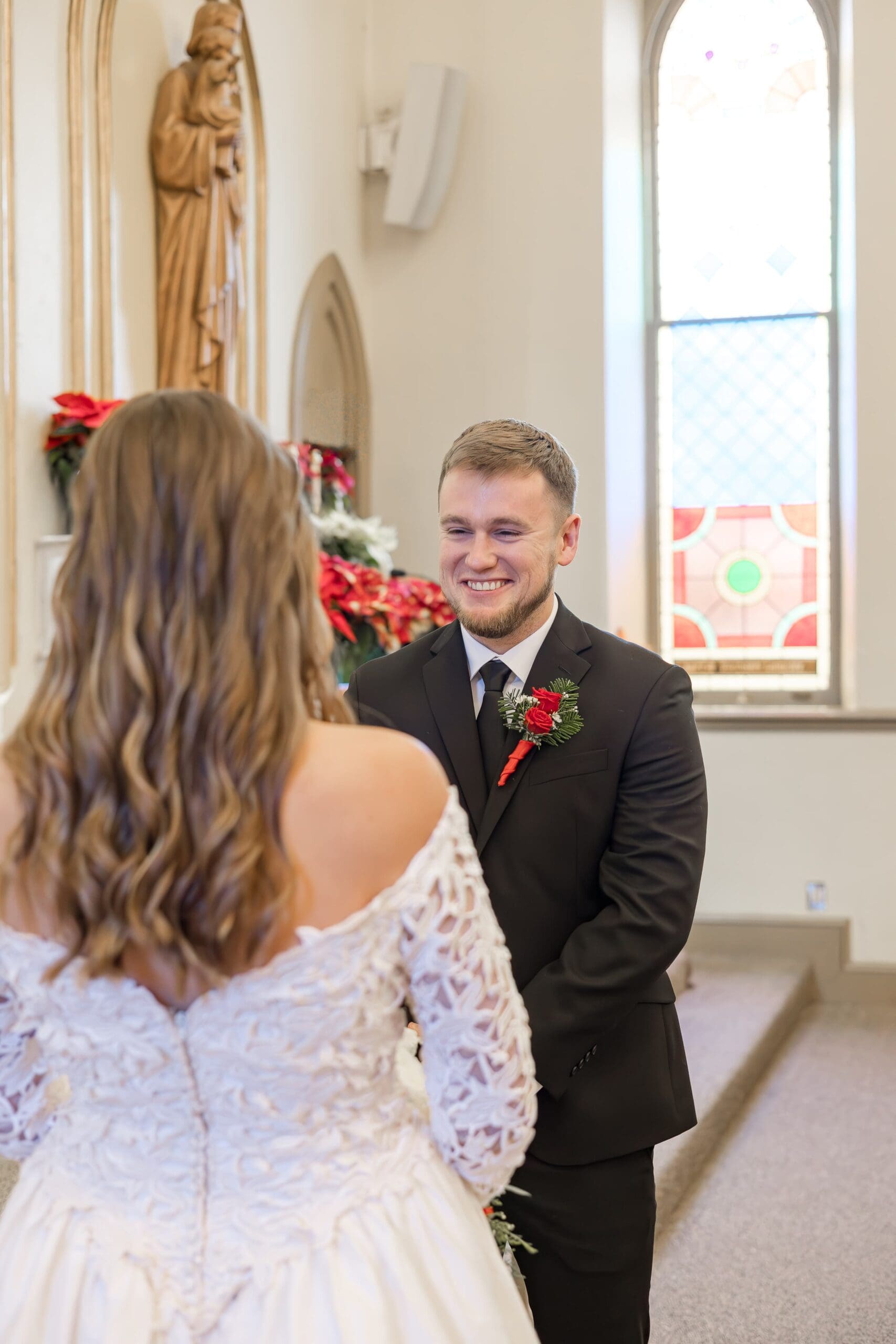 man at the alter stands in front of his bride smiling at her in a black suit and red boutonniere.