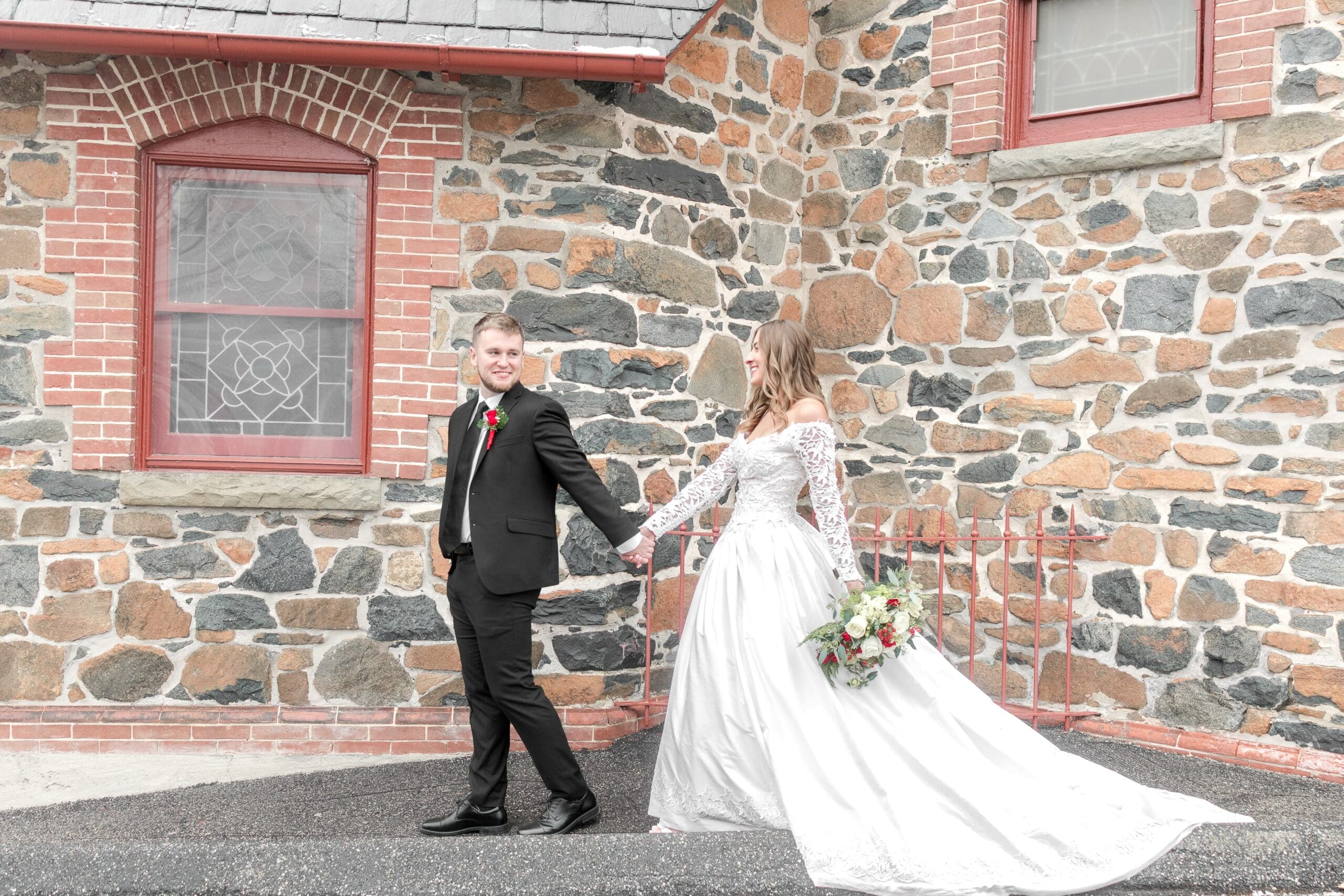 Beautiful bride in a white lace dress being guided down a black walkway by her husband in a black tuxedo out front of their ceremony building, a Catholic church.
