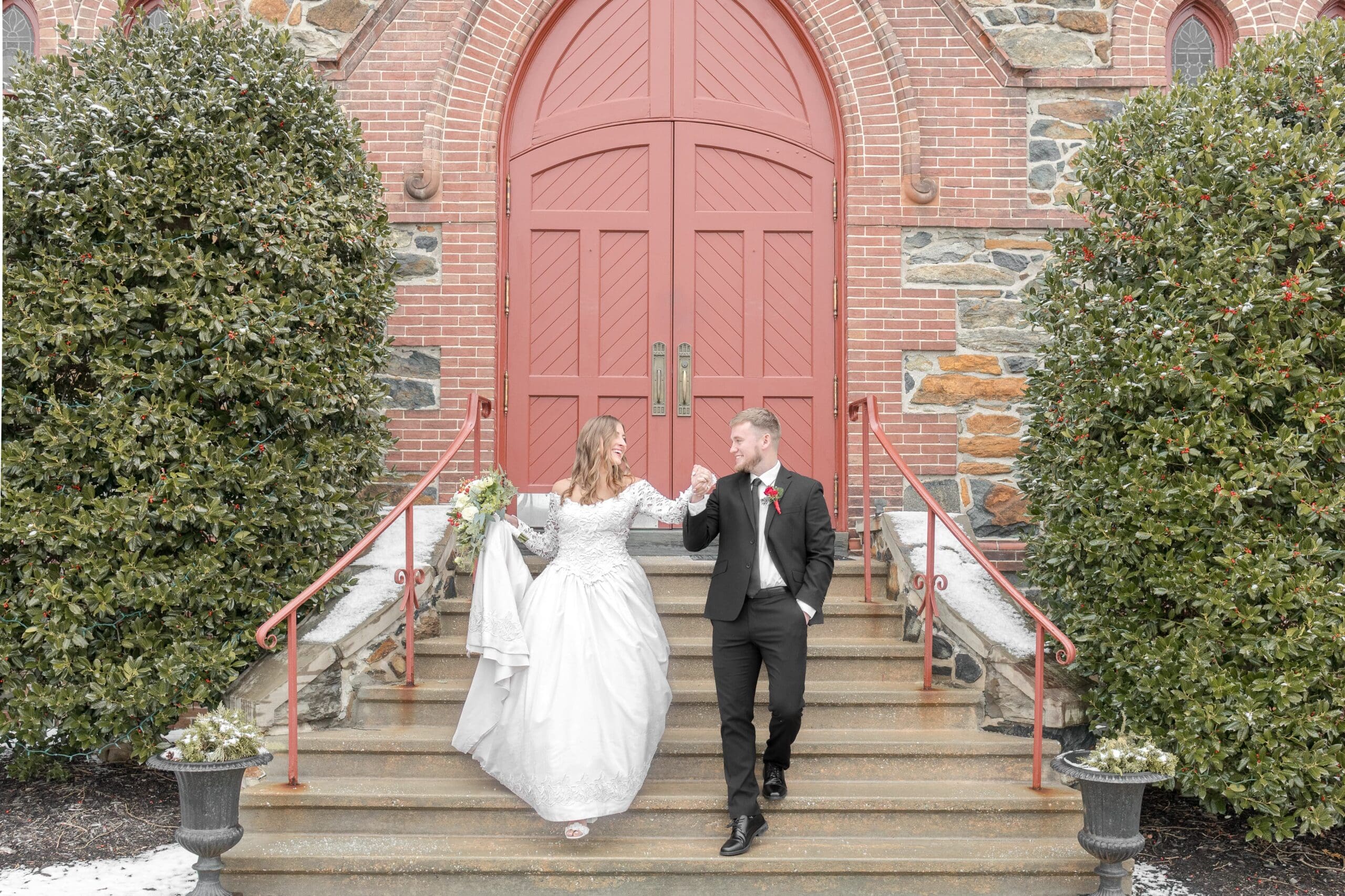 Beautiful bride in a white lace dress being guided down the stairs by her husband in a black tuxedo out front of their ceremony building, a Catholic church.