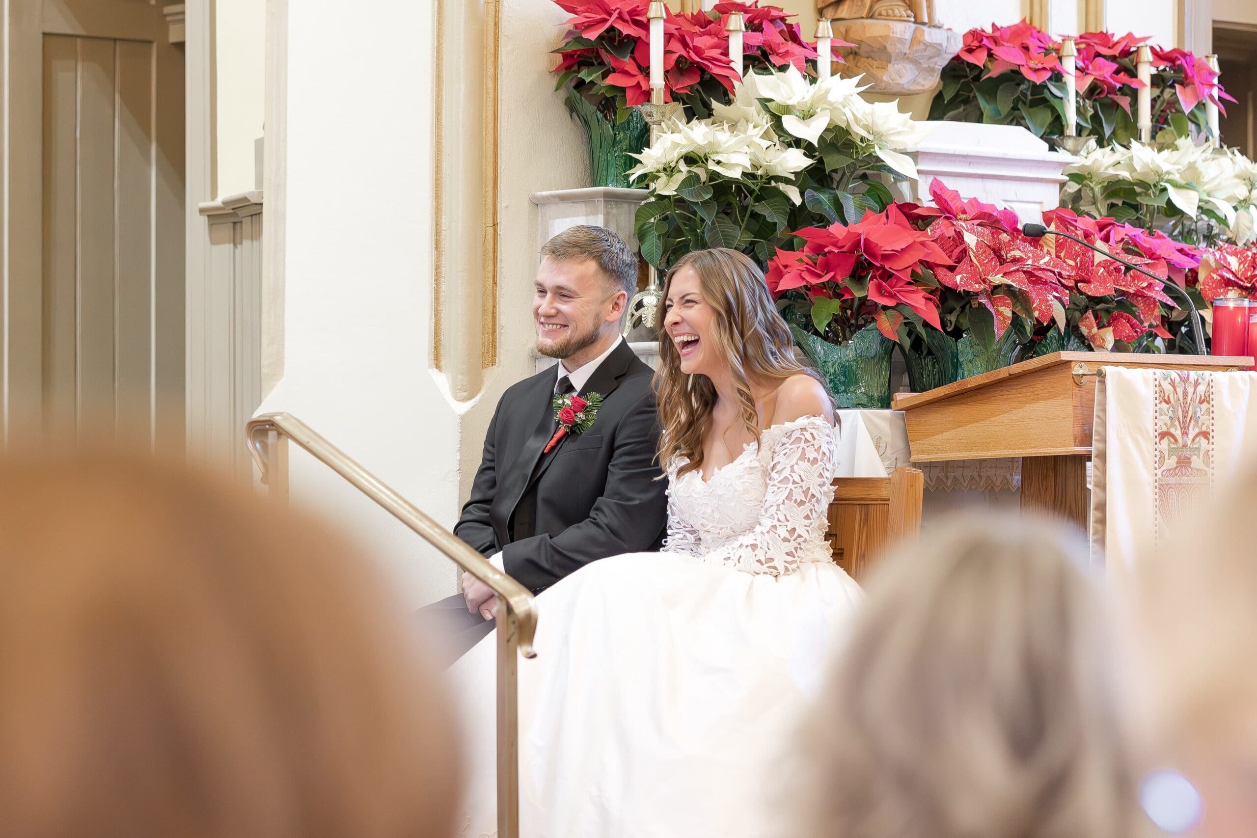 Beautiful bride in a white lace dress and groom in black suit sitting and smiling. They are surrounded by poinsettia flowers and guests' heads are in the foreground/