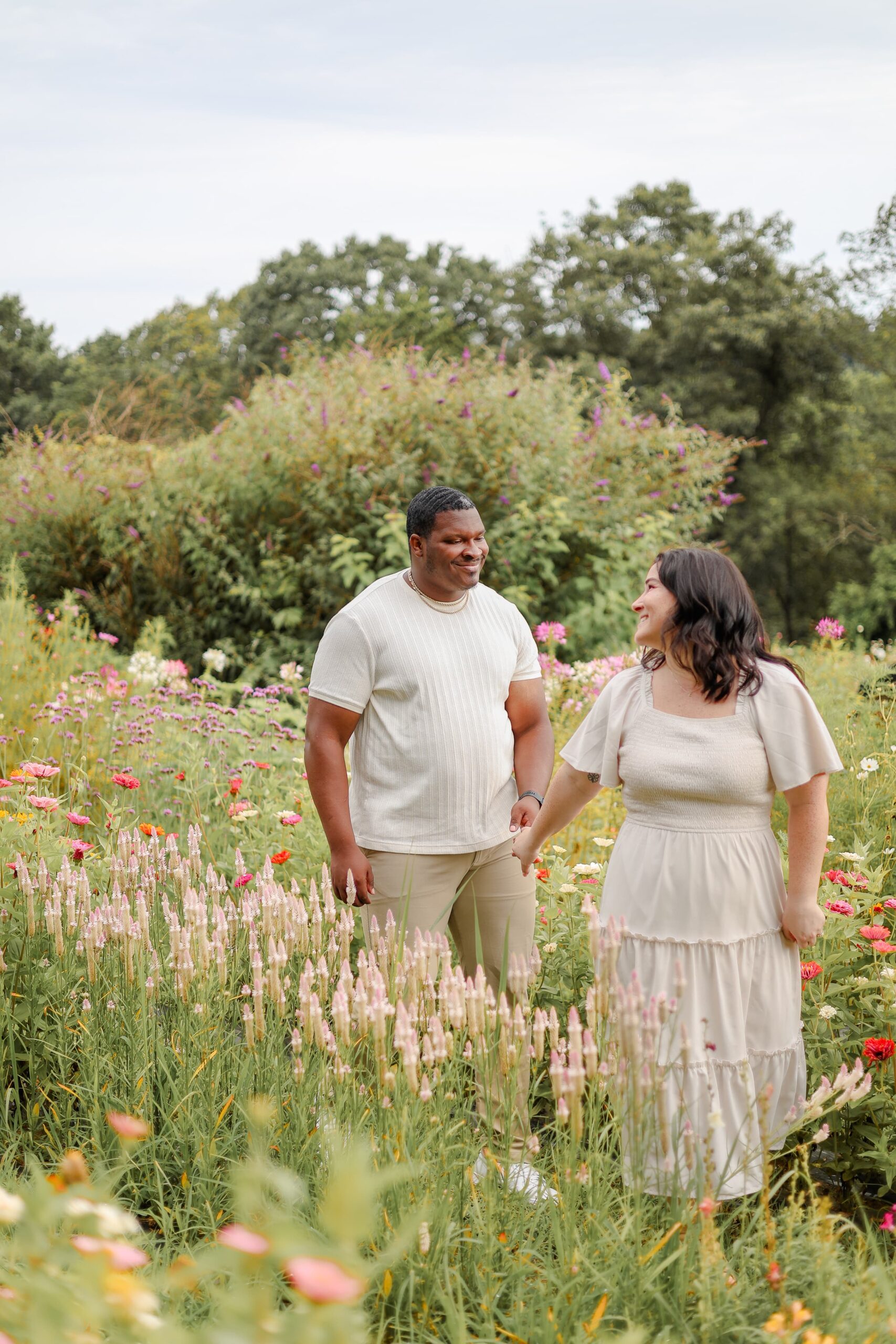 Man and woman walking in a wildflower field as the woman in an ivory dress looks back at her fiance. They are holding hands as he is smiling at her.