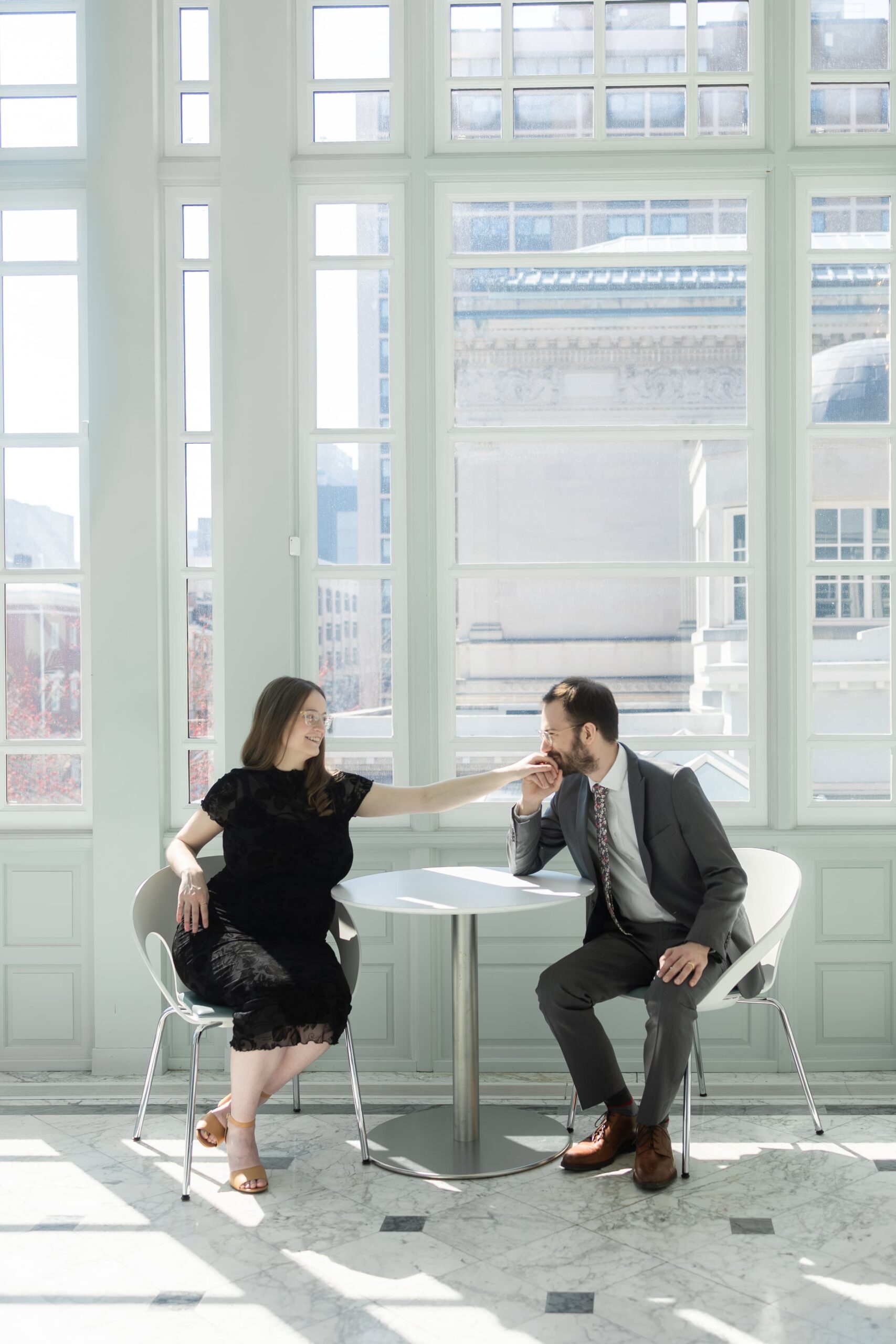 Husband and wife sit at a table at the Walters Art Museum. The husband in a suit and tie, kisses his wife's hand from across the table.